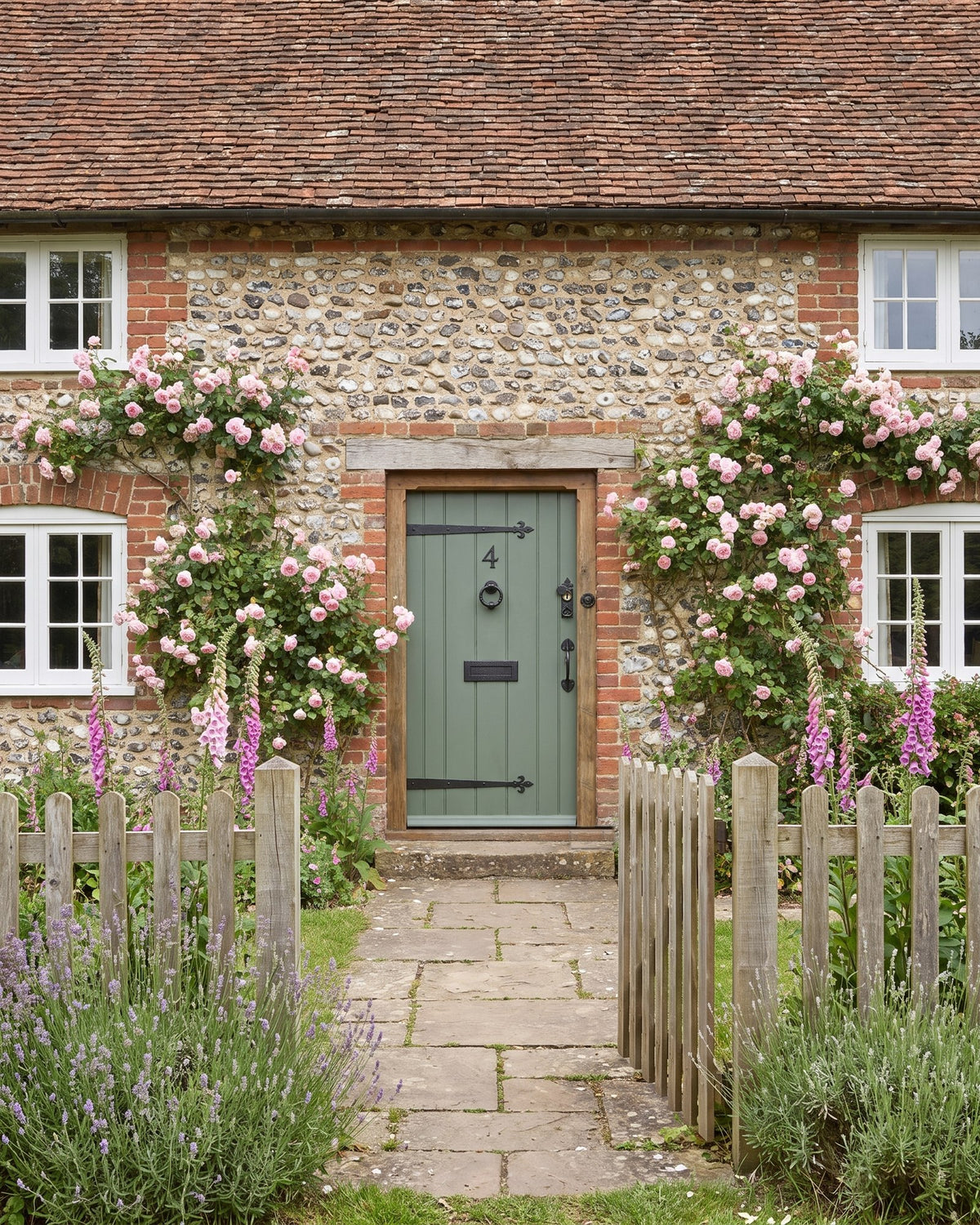 Brick and flint cottage with a clay tile roof, white casement windows and pink climbing roses around a grey-green boarded front door, seen from a picket gate