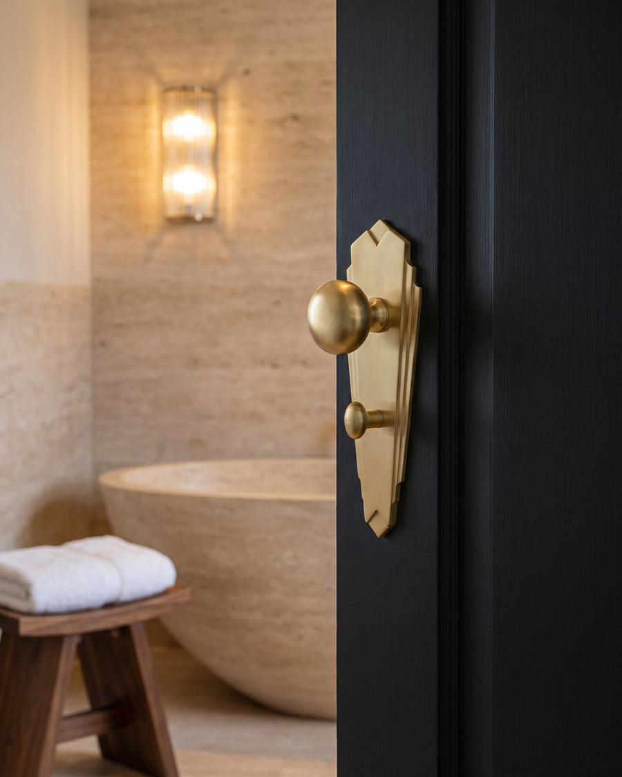 Satin brass Broadway knob on a bathroom plate with turn on a dark en-suite door, travertine walls and stone bath beyond