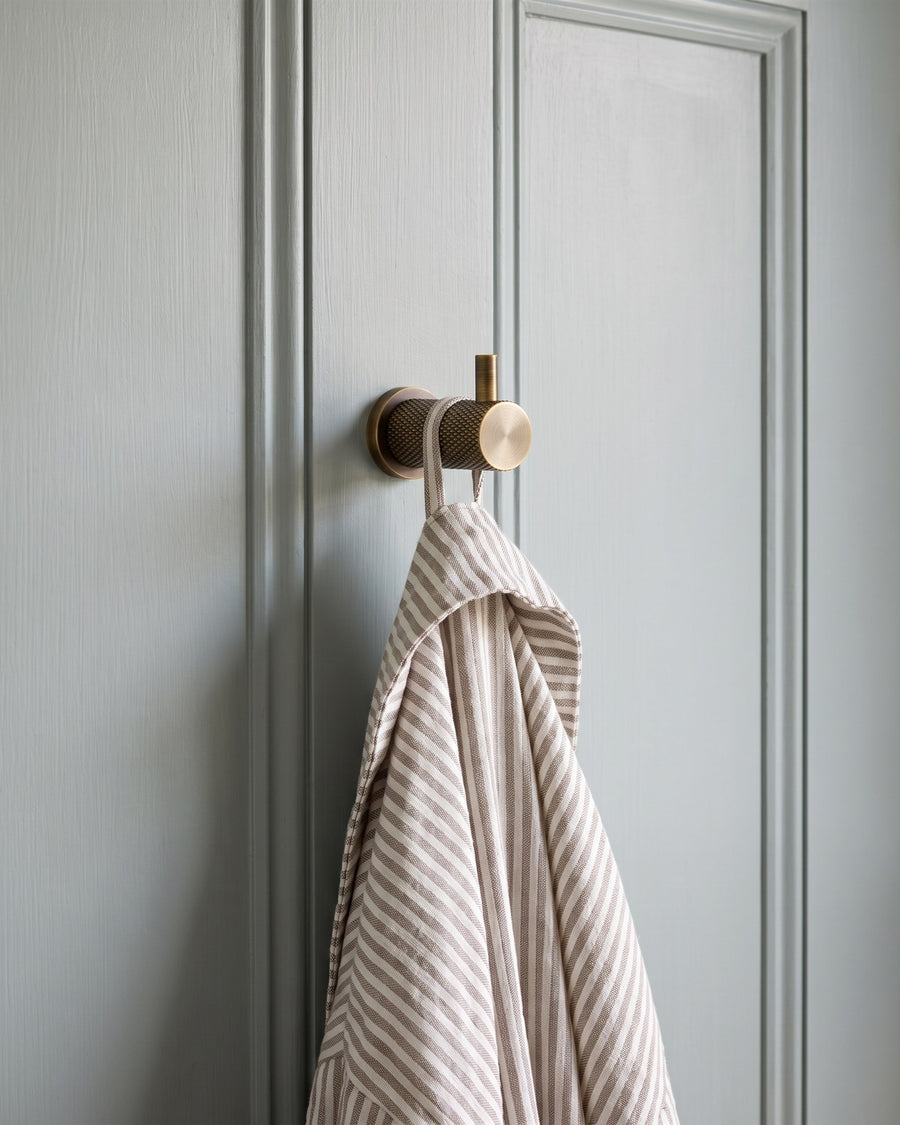 Knurled antique brass Brunel robe hook holding a striped linen bathrobe on a grey-blue panelled door