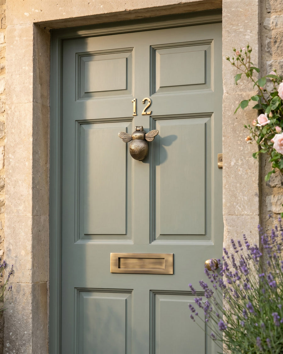 Grey-green front door in a Cotswold stone cottage with an antique brass bumble bee knocker, brass house numbers and letter plate, lavender and roses beside