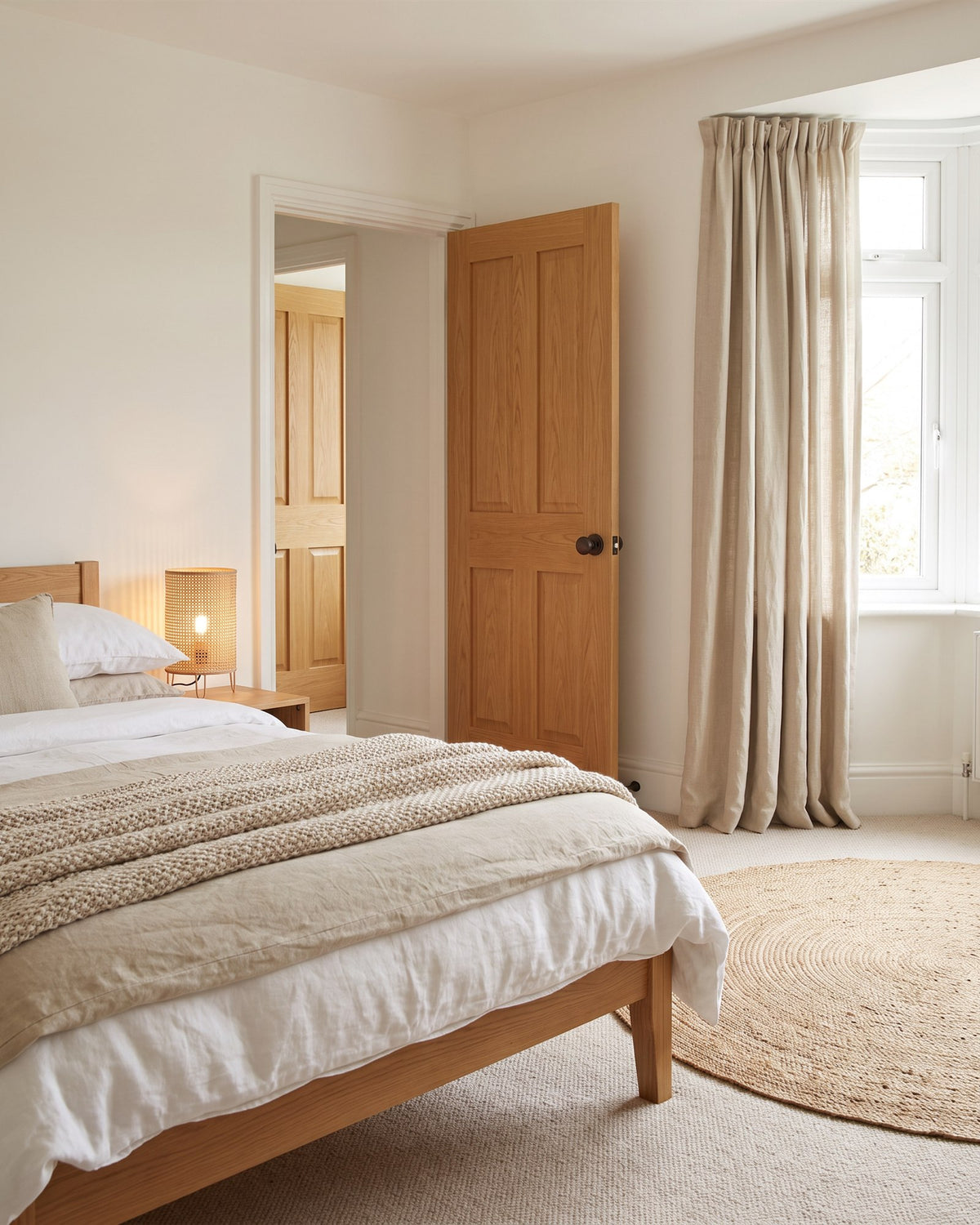 Calm bedroom with an oak bed, sand and white linen, a jute rug and linen curtains, a golden oak door with a matt bronze knob