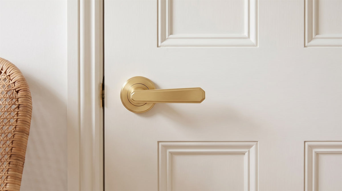 Century satin brass lever on a round rose on a warm white four-panel door, with a rattan chair beside the frame