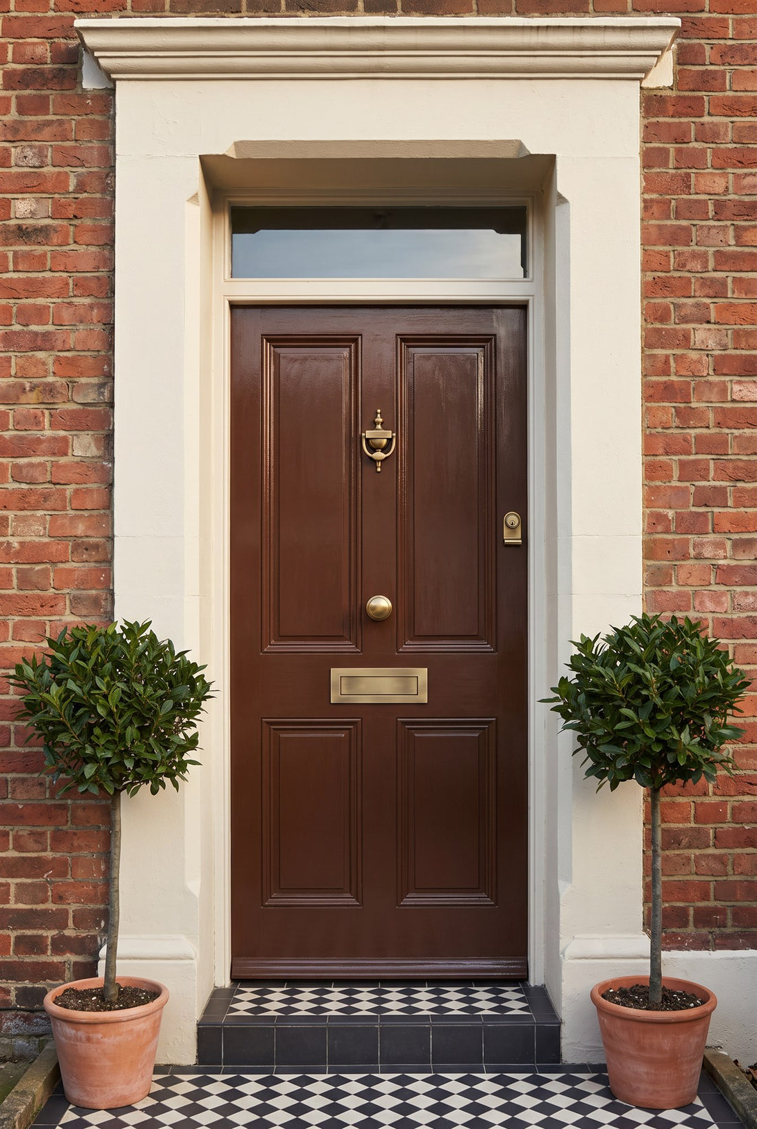 Glossy chocolate brown four-panel Edwardian front door in a cream doorcase with a chequered step and bay trees, fitted with an antique brass urn knocker, centre knob, letter box and cylinder pull