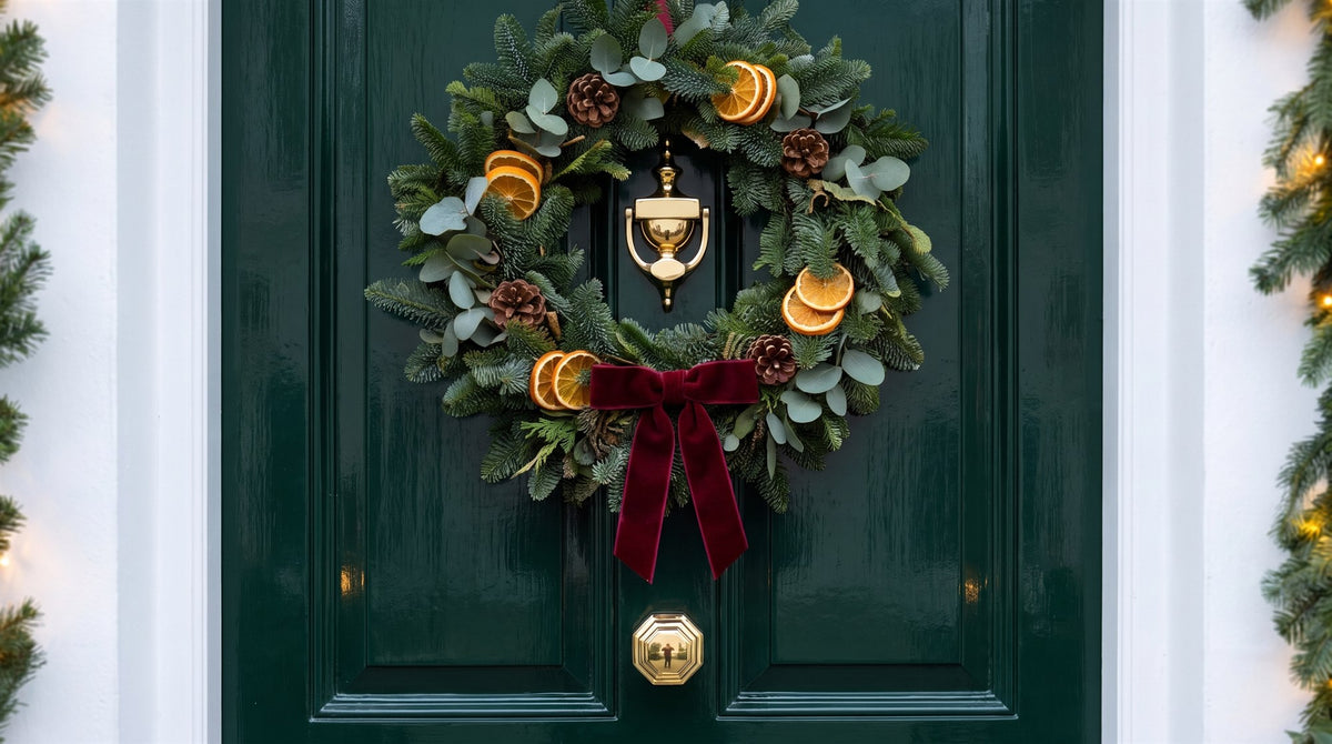 Polished brass urn knocker framed by a fir, eucalyptus and dried orange wreath with a red velvet bow on a glossy bottle-green front door, an octagonal brass centre knob below