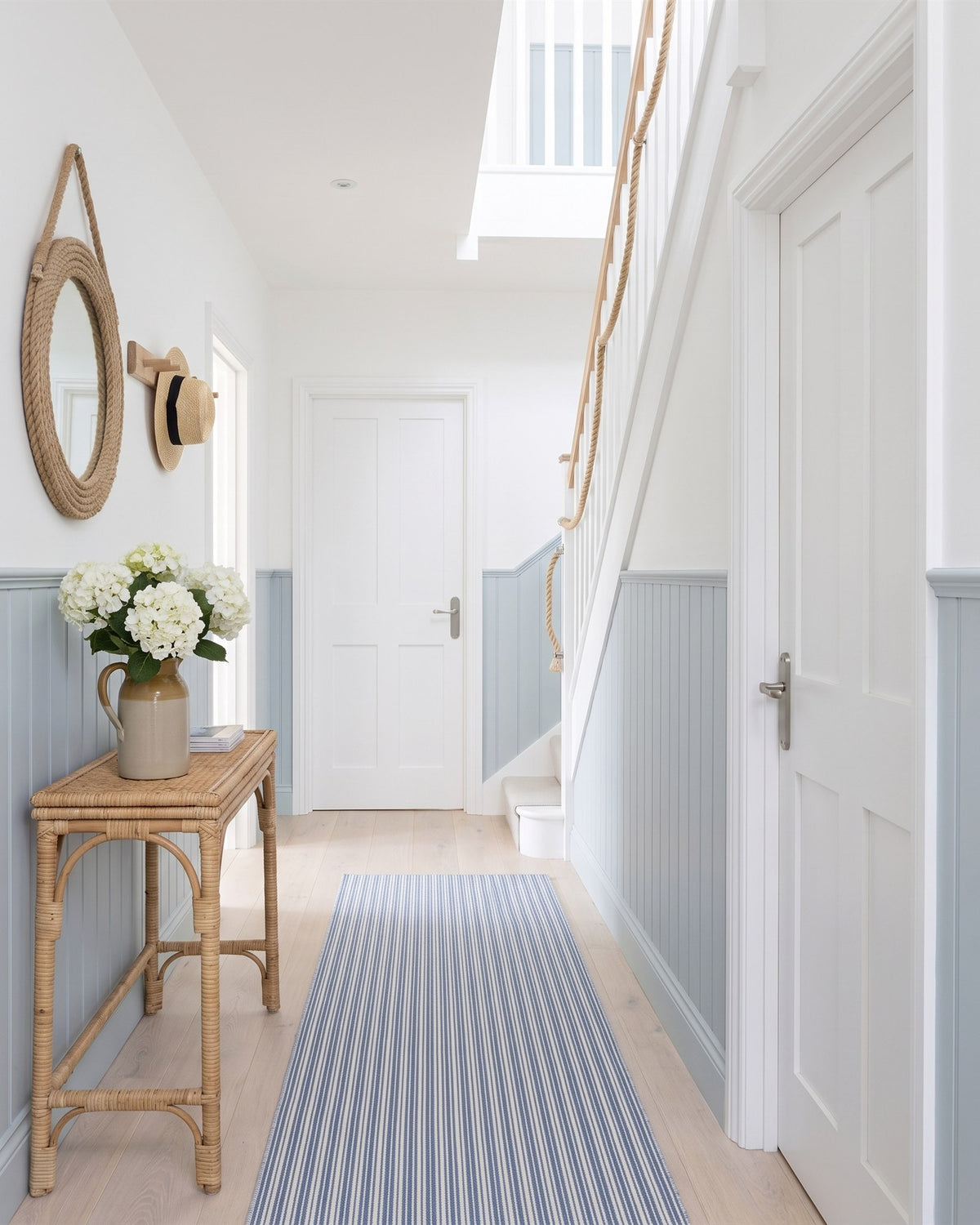 Bright coastal hallway with powder blue tongue-and-groove panelling, white Shaker doors with satin nickel lever handles, a striped runner and a rattan console