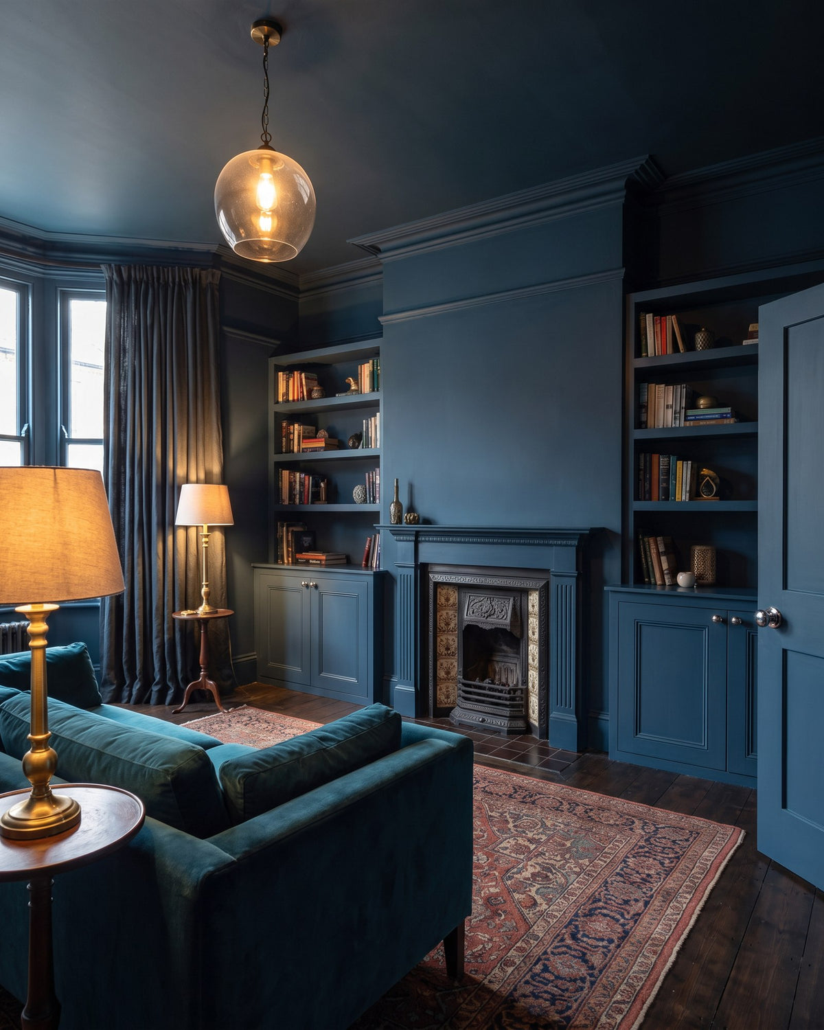 Edwardian living room painted ink blue on walls, woodwork and ceiling, with alcove shelves, a cast iron fireplace, teal velvet sofa and brass lamps