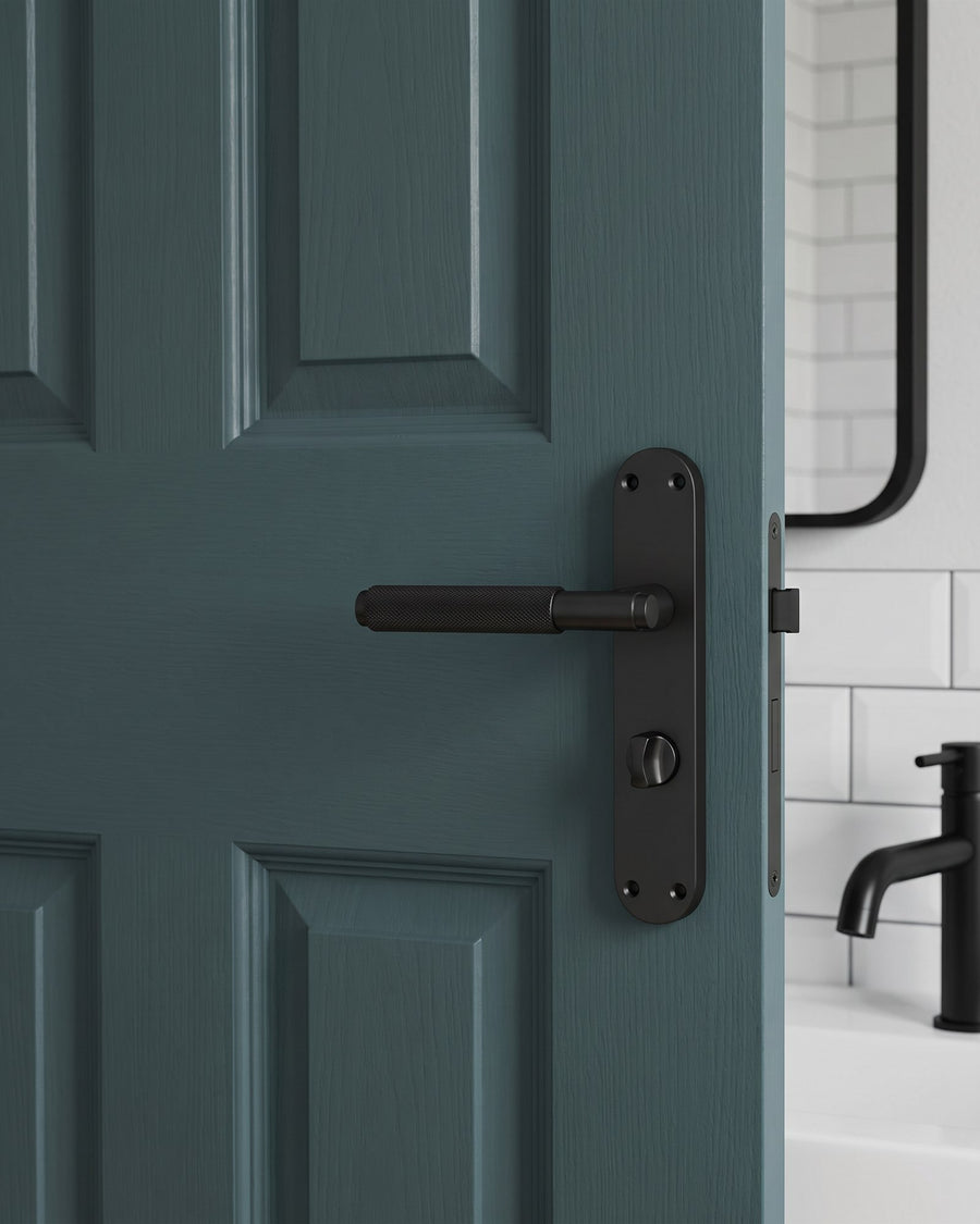 Matt black Connaught knurled bathroom handle with thumbturn on a deep green panelled door, white metro tiles and a black tap beyond