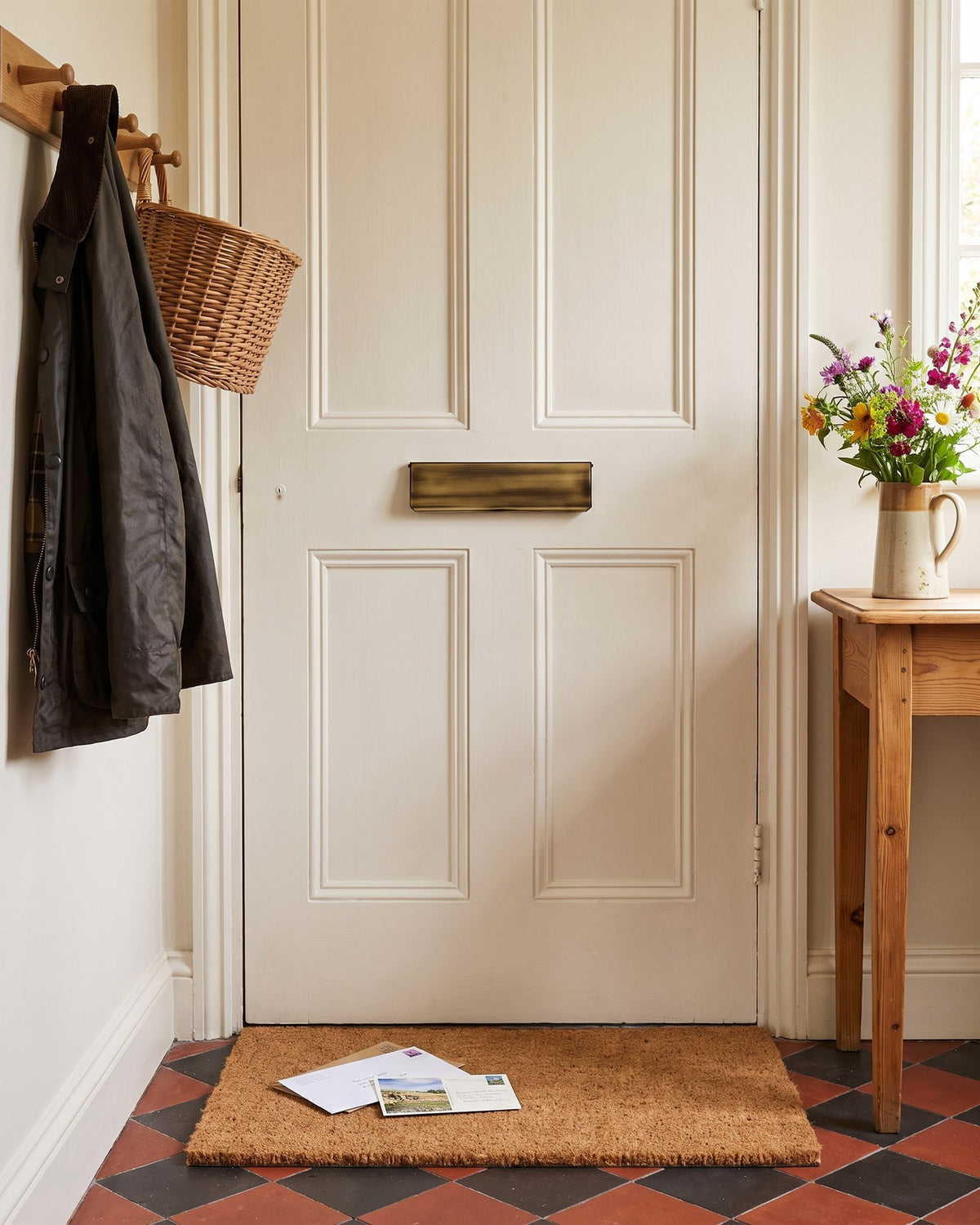 Cottage hallway with red and black quarry tiles, letters and a postcard on a coir mat below an antique brass interior flap, a waxed jacket on a peg rail and garden flowers in a jug