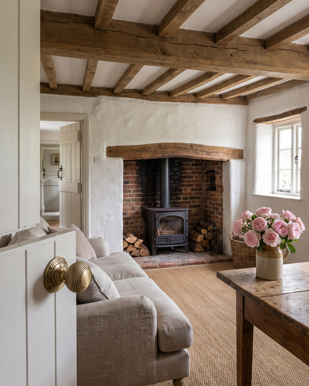 Antique brass reeded knob on an off-white cottage door opening onto a beamed sitting room with a brick inglenook and woodburner