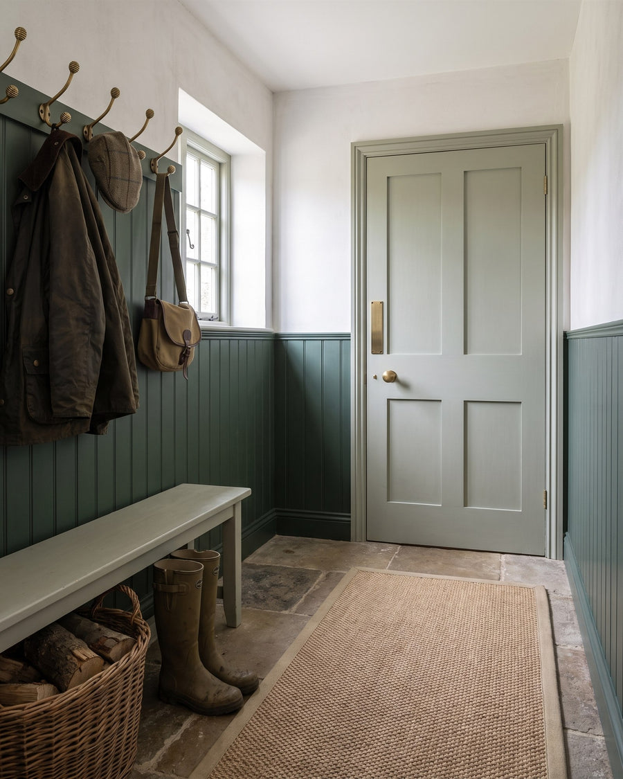 Country house boot room with green tongue-and-groove panelling, a row of brass beehive coat hooks, a sisal runner on flagstones and a grey-green door with a beehive knob