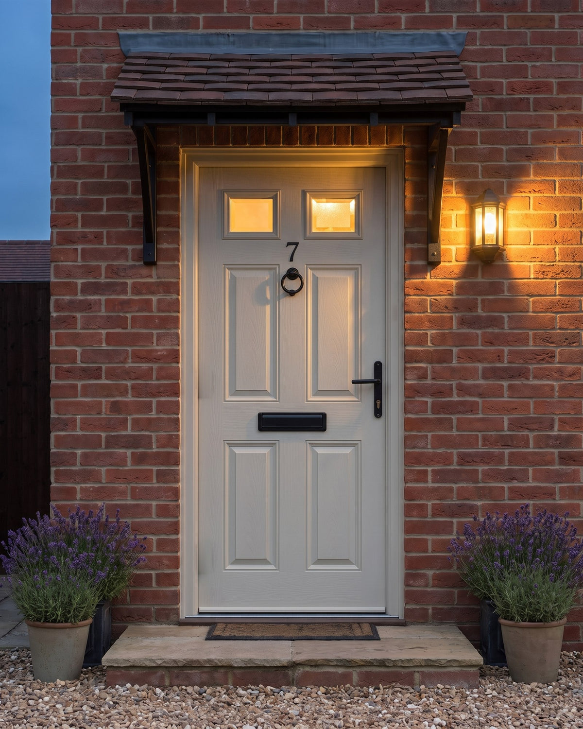 Cream composite front door at dusk with a glowing black lantern and lit glass panes, the antique black knocker, lever and letter plate picked out
