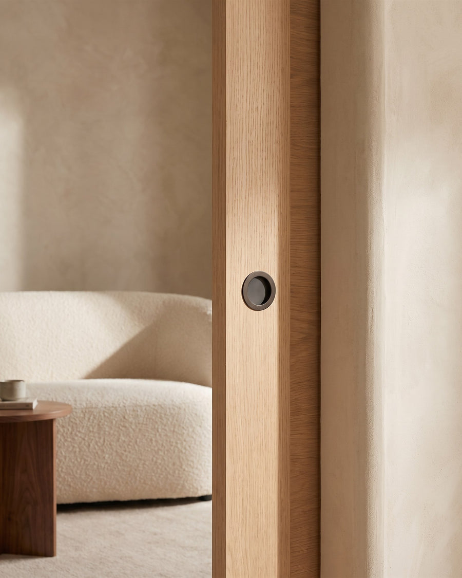 Pale oak pocket sliding door with a dark bronze circular flush pull, opening onto a snug with a boucle sofa