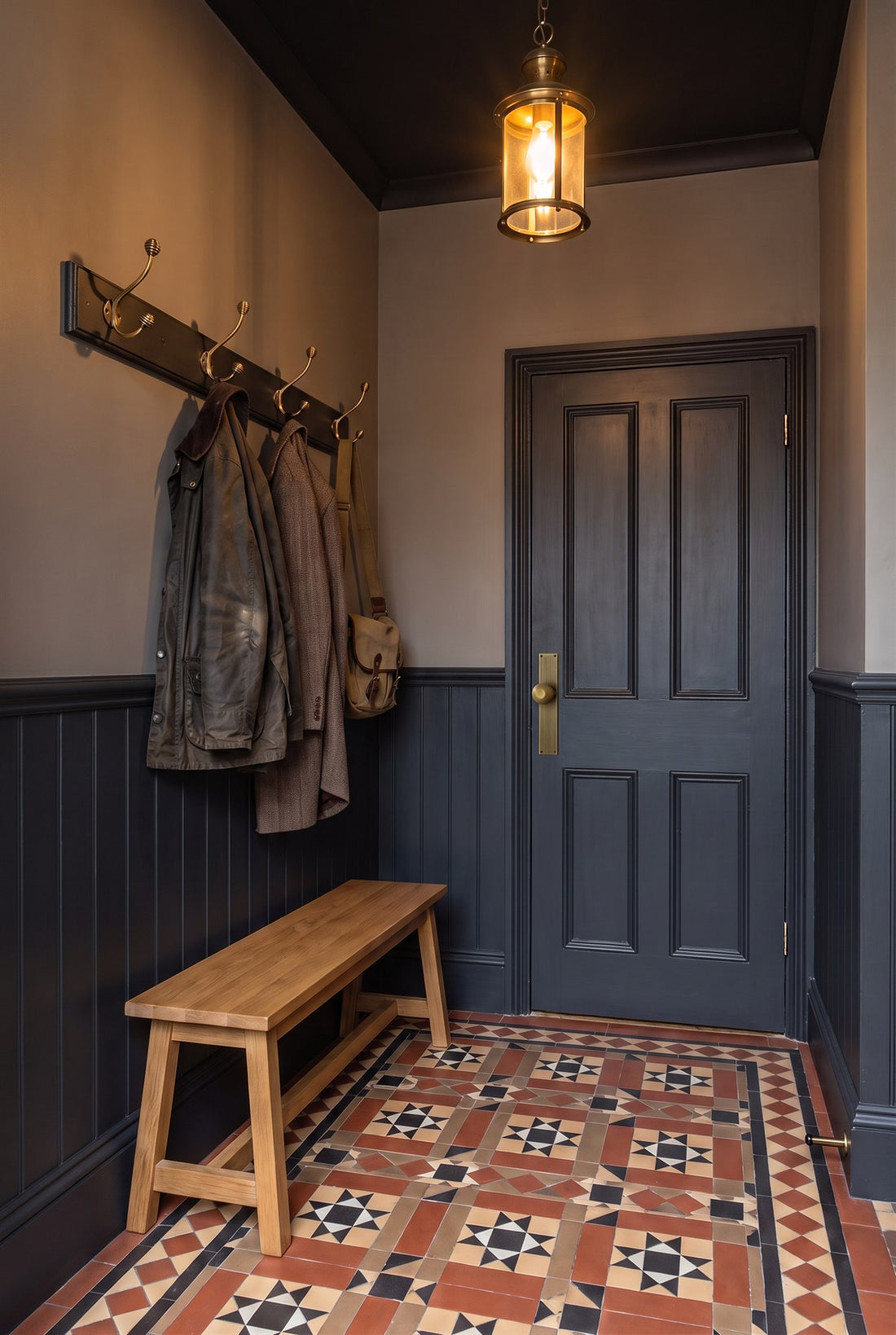 Victorian hall with off-black panelling and encaustic tiles, a bench under aged brass beehive coat hooks, and an off-black door with an antique brass beehive knob on a plate and a projection door stop