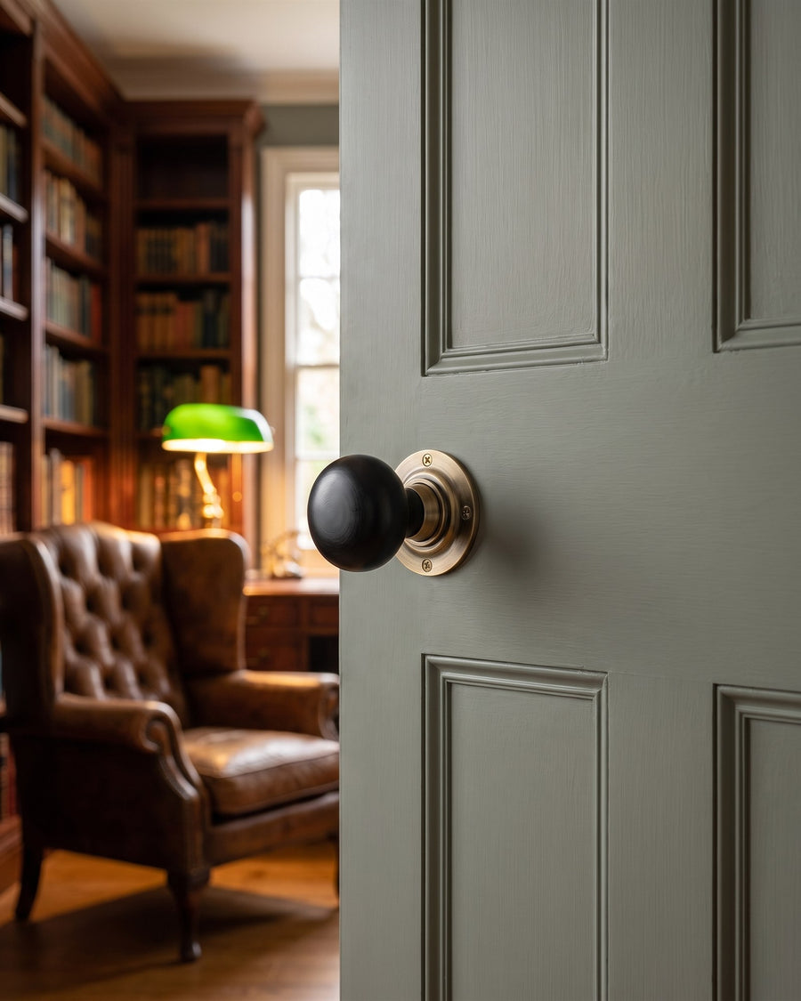 Black ebony mushroom knob on an antique brass rose on a grey-green study door, leather wing chair and green banker's lamp beyond