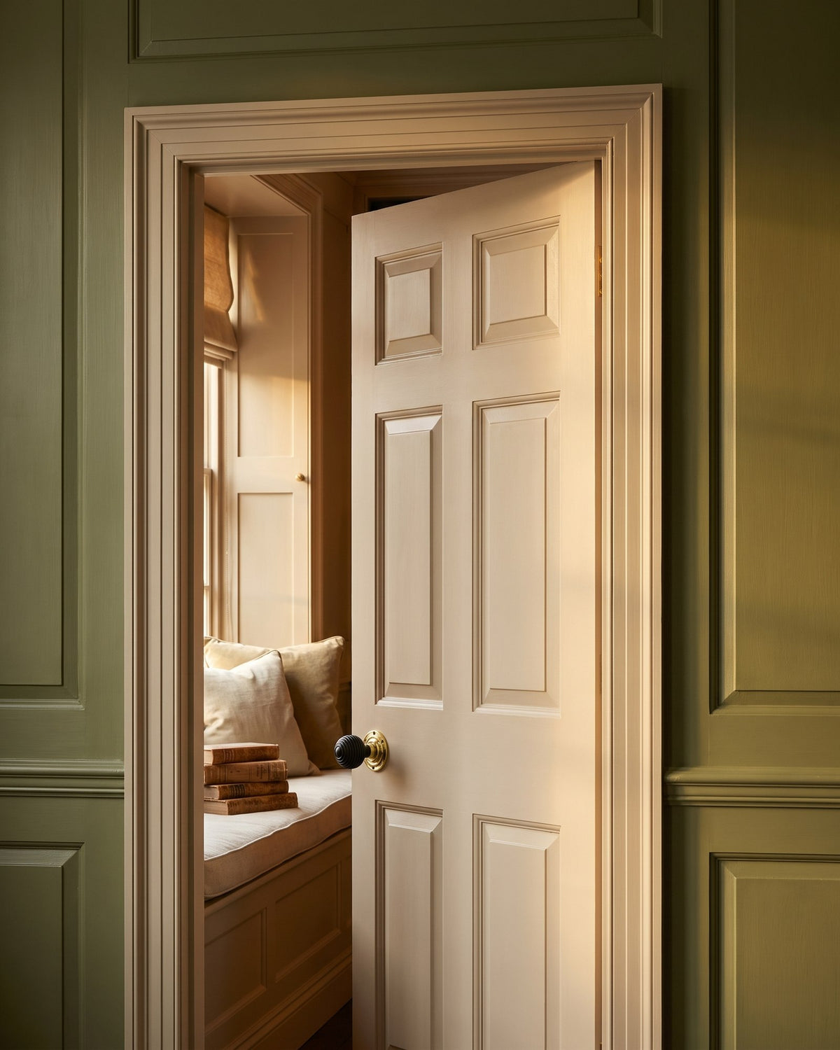 Stone coloured door with an ebony and polished brass reeded knob open to a reading room window seat, olive panelling around