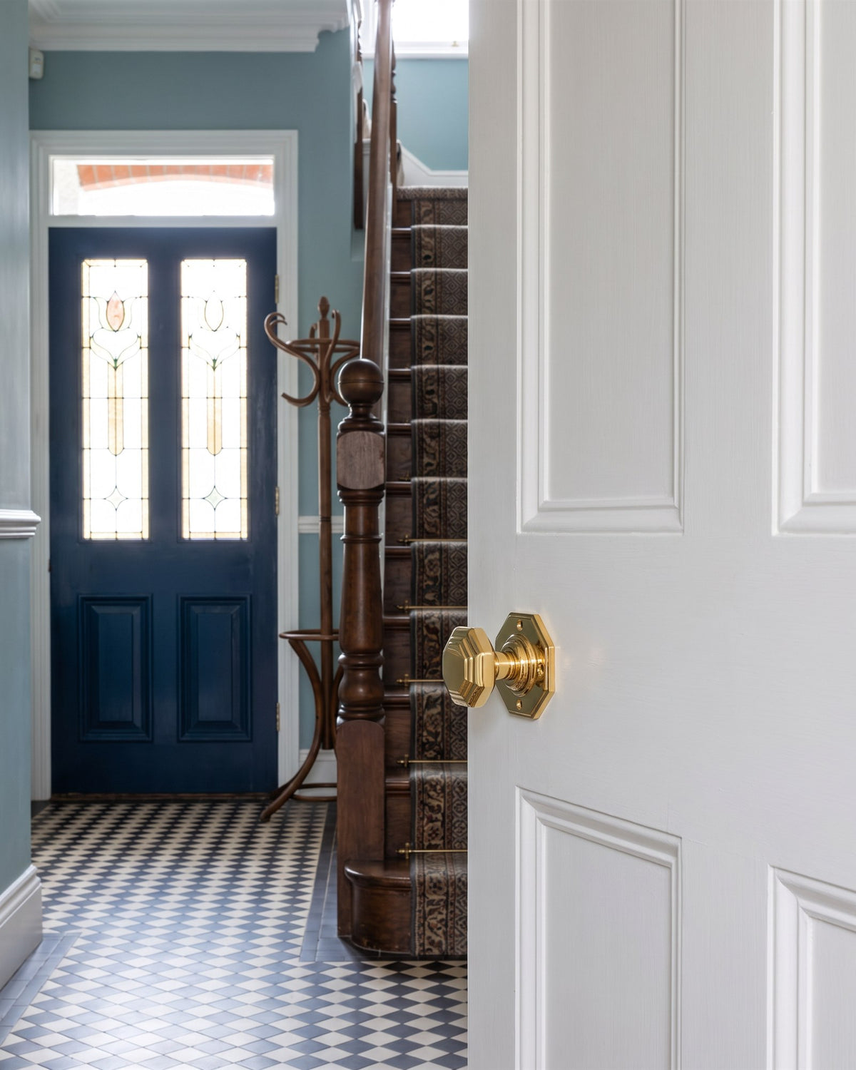 Polished brass octagonal knob on a white panelled door in an Edwardian hall with a chequerboard floor, oak staircase and the blue stained-glass front door beyond