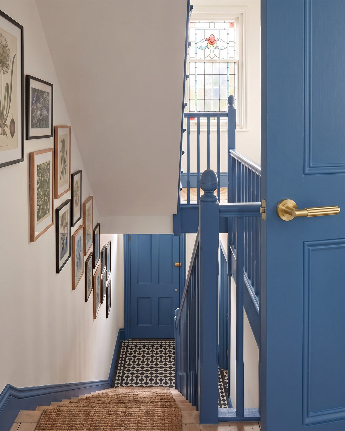 Edwardian stairwell with mid blue spindles, skirting and doors against warm white walls, beside a blue door with a satin brass Bloom lever