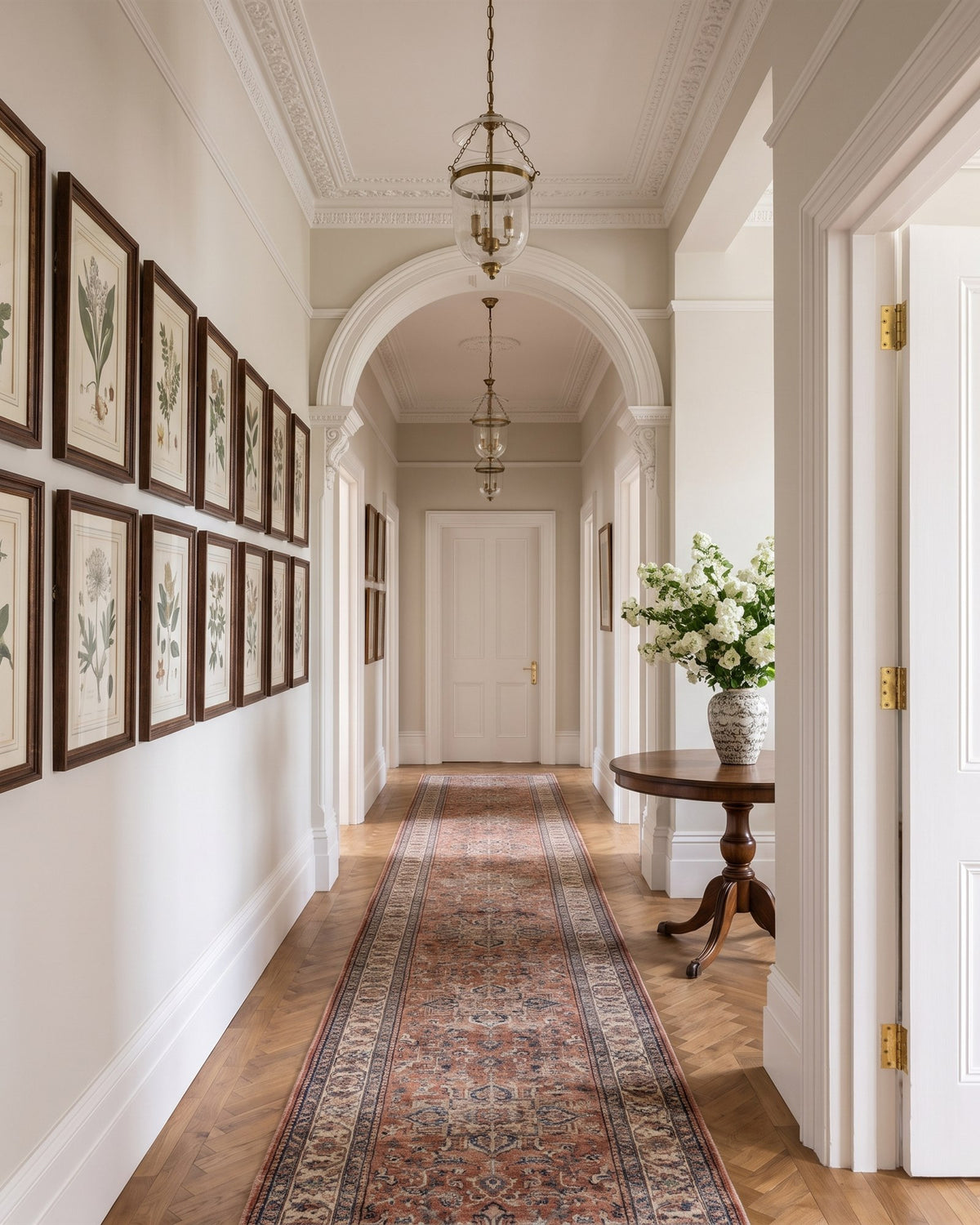 Long entrance hall of an Edwardian mansion flat with herringbone parquet, a Persian runner, botanical prints, an arch and glass lanterns