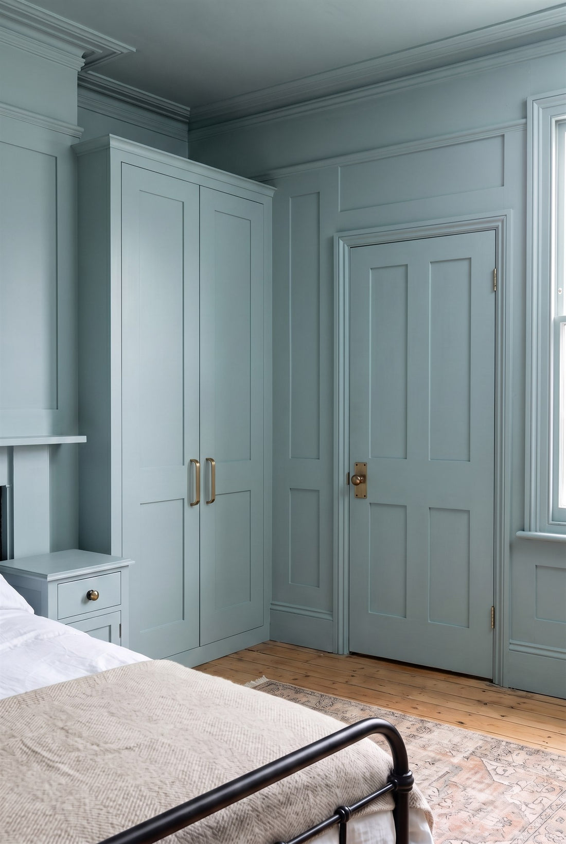 Edwardian bedroom with full-height blue-green panelling, built-in wardrobes with antique brass D handles, a panelled door with an antique brass knob on a latch plate, an iron bed and a bedside table with a brass knob
