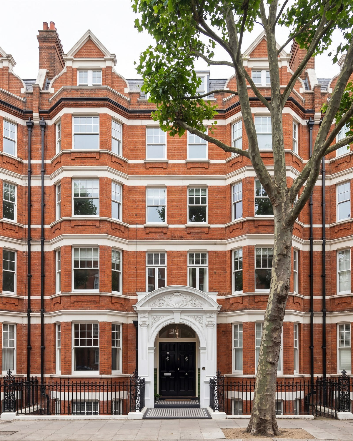Red-brick Edwardian mansion block with white stone bands, bay windows and an ornate arched entrance behind black railings, a plane tree on the pavement