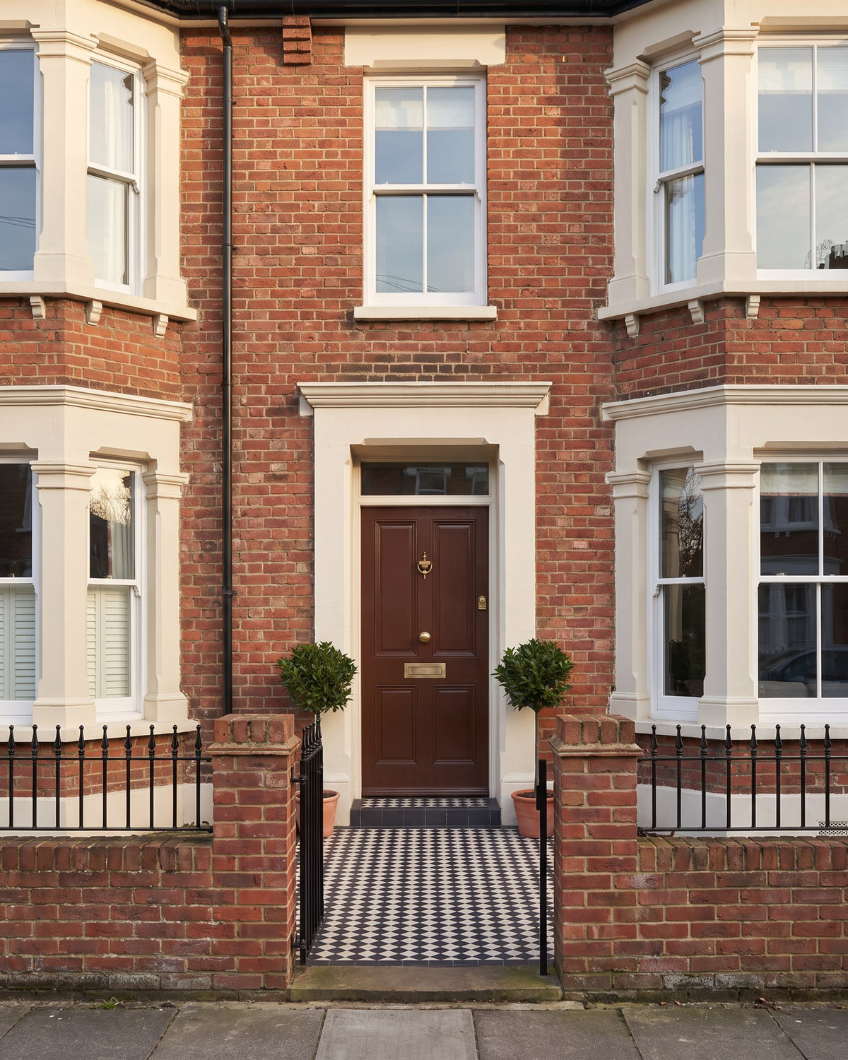 Edwardian red-brick terrace with cream bay windows and doorcases, the centre house with a glossy chocolate brown front door, bay trees and a chequered path