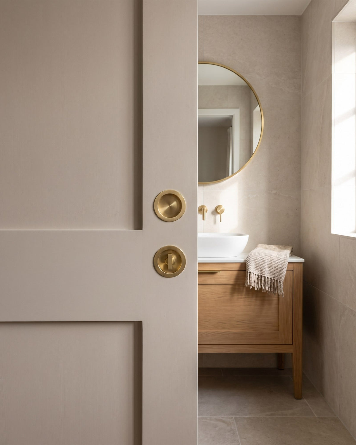 En suite pocket door from the bathroom side with a circular satin brass flush pull above a matching circular turn, stone tiles and an oak vanity beyond