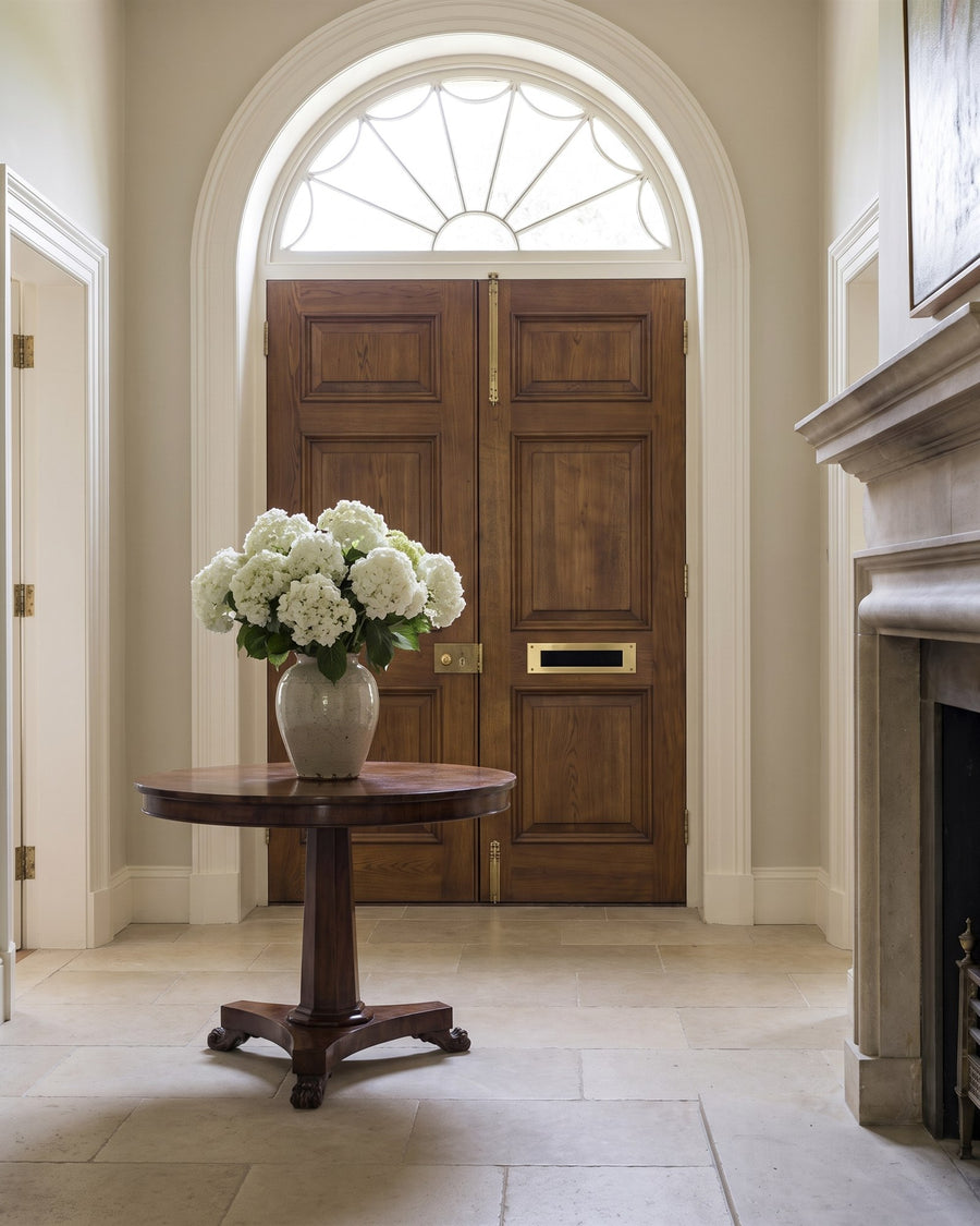 Stone-floored entrance hall with a round mahogany table of white hydrangeas in front of the inside of tall oak double doors and a semicircular fanlight