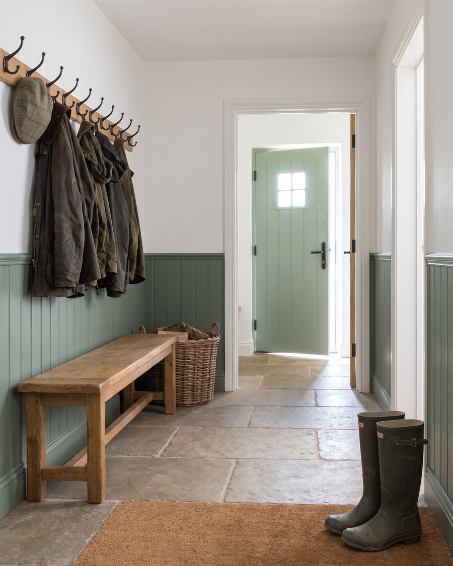 Farmhouse boot room with flagstones, an oak bench, black coat hooks and wellies, the green front door seen through the doorway