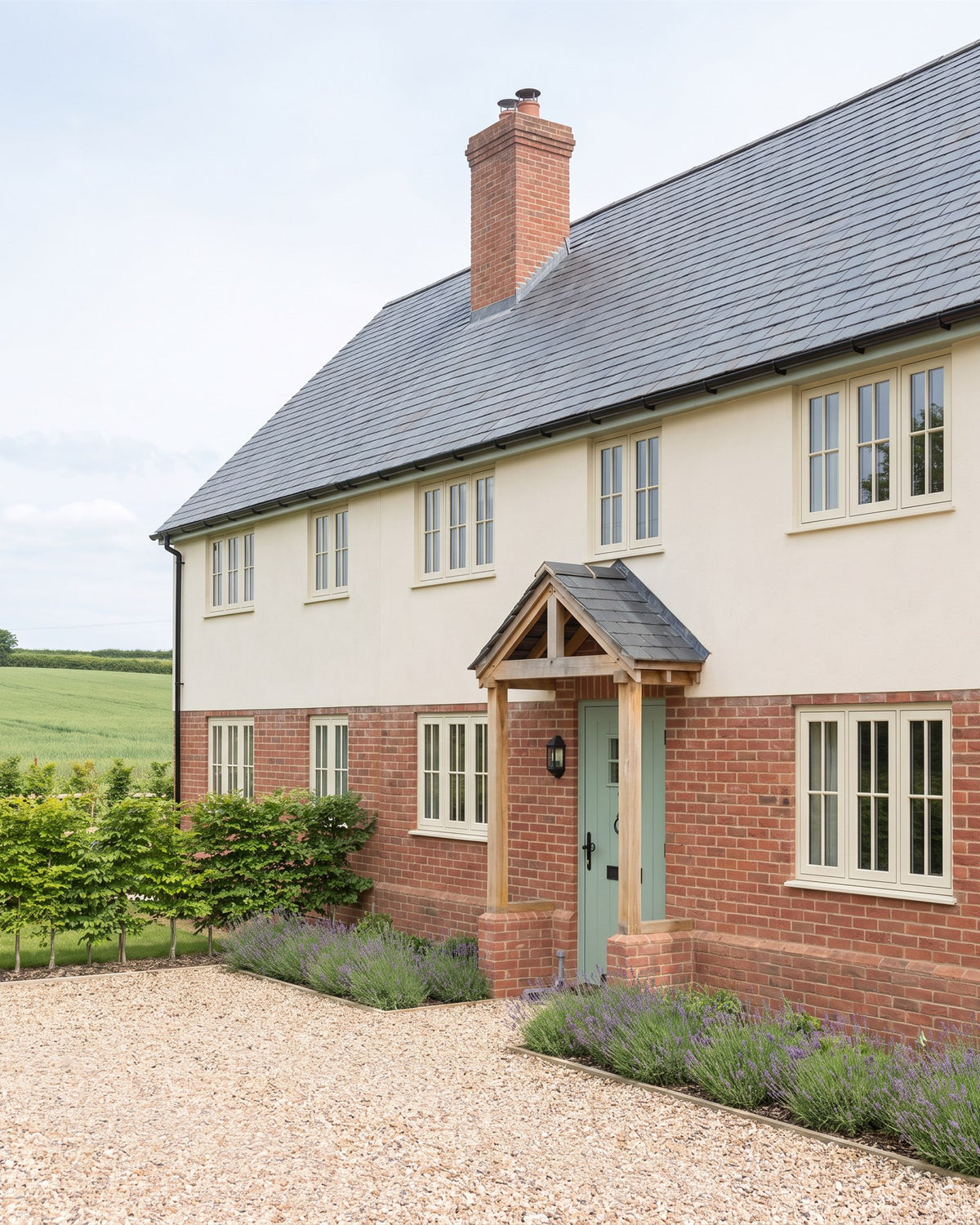 Farmhouse-style new build in red brick and lime render with a slate roof, cream casement windows and an oak porch over a green front door, gravel and lavender