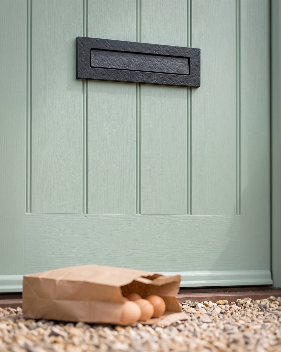 Forged black hammered architectural letter plate on a heritage green composite door, a paper bag of farm eggs on the gravel below