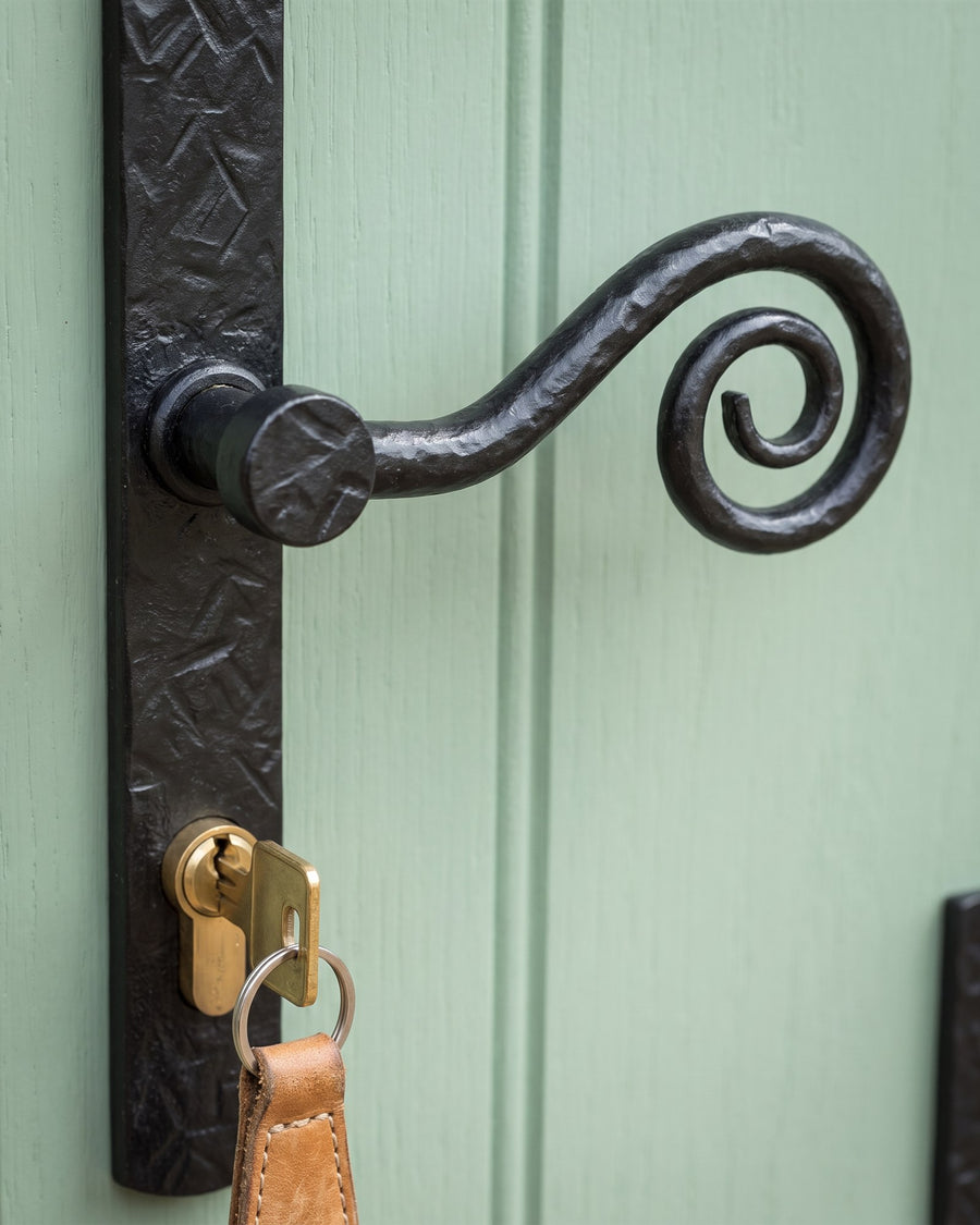 Forged black monkey tail lever curling into a spiral above a brass euro cylinder with a key and tan leather fob