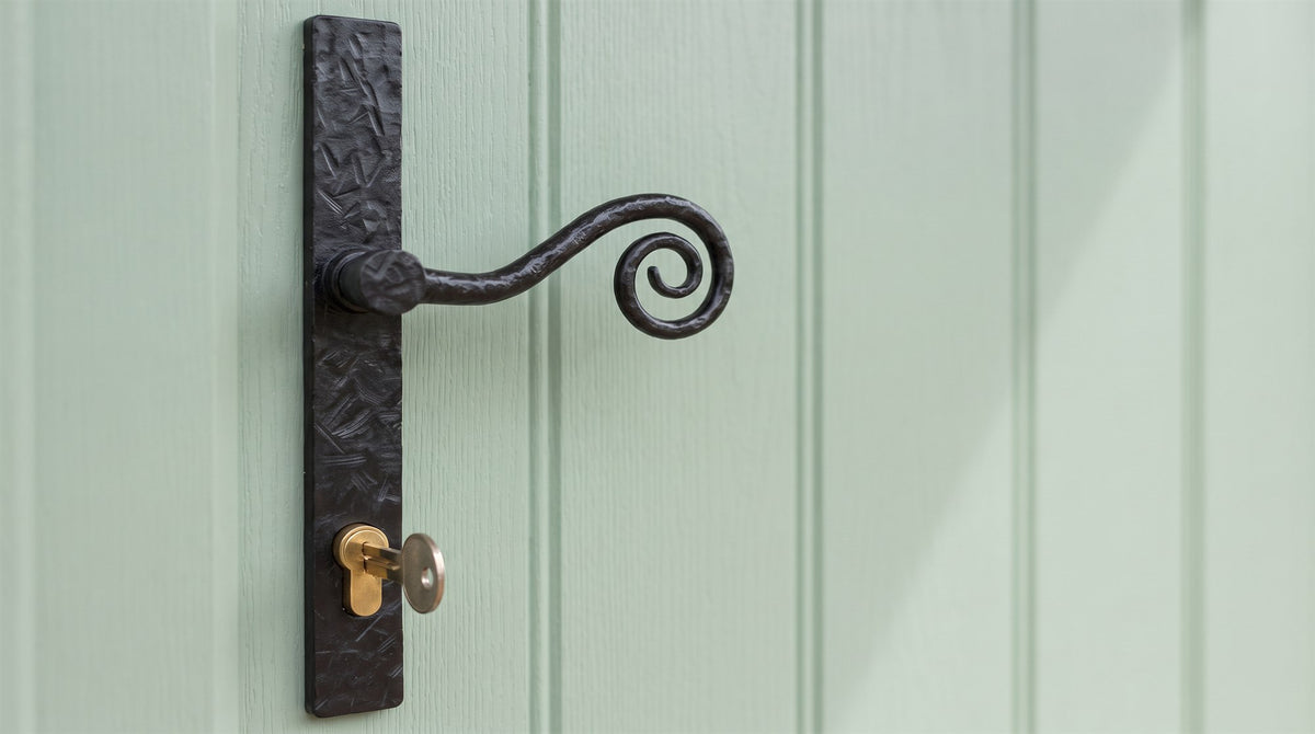 Forged black monkey tail lever with a spiral curl on a long hammered backplate, on a heritage green boarded composite door