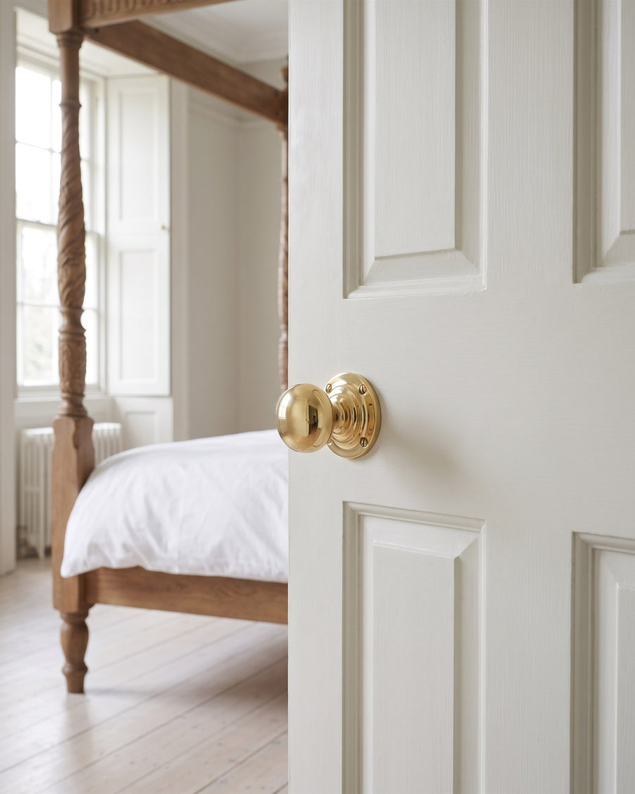 Plain round polished brass door knob on a chalky white six-panel bedroom door, four-poster bed beyond