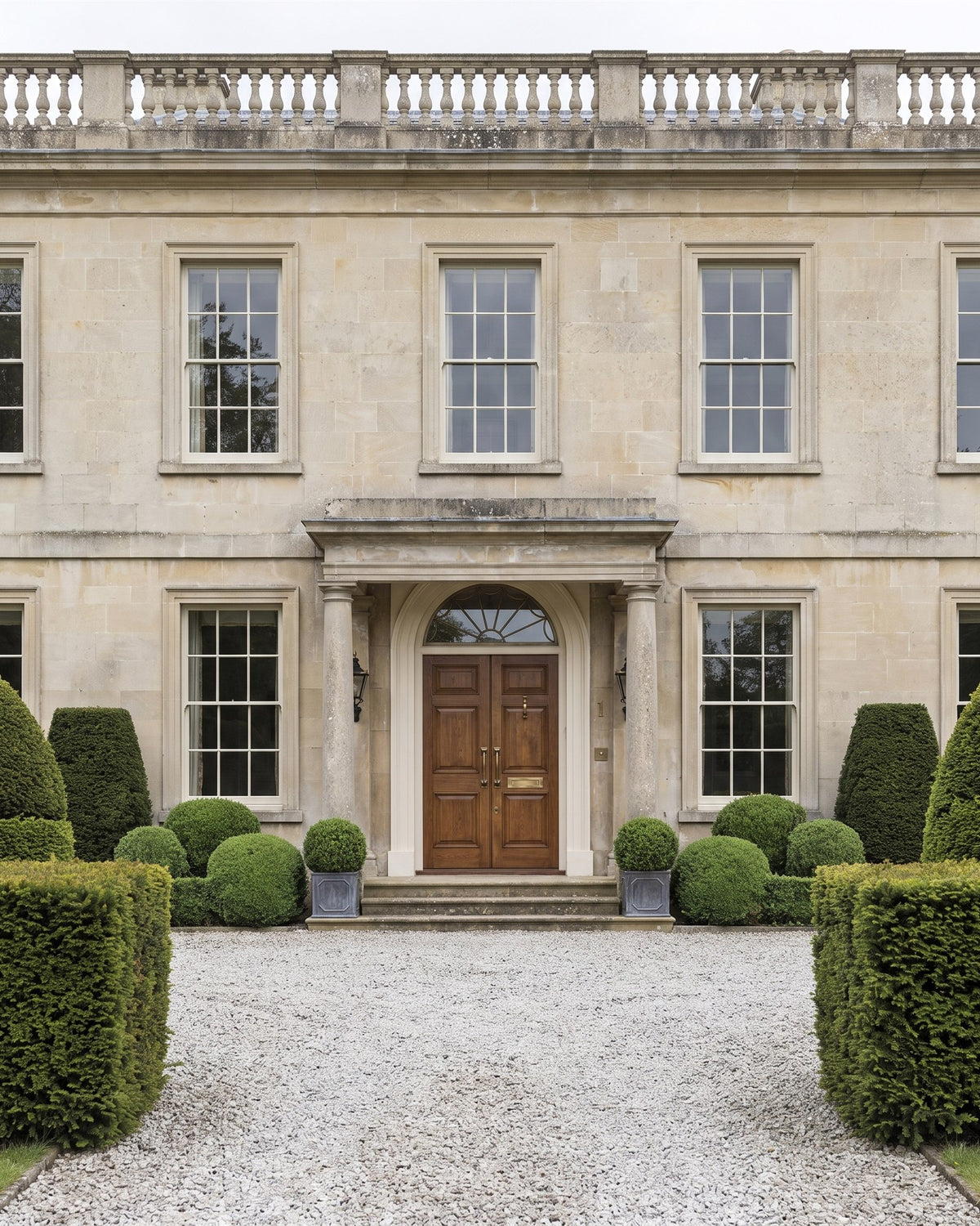 Symmetrical Bath stone Georgian country house with sash windows, a balustraded parapet and a columned portico around oak double doors, seen from a gravel drive lined with yew and box