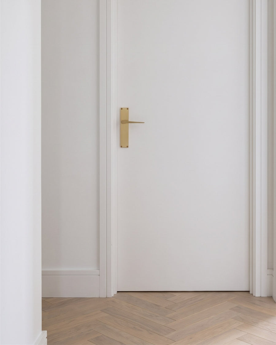 Gio satin brass lever on a slim backplate on a white flush internal door above a pale oak herringbone floor