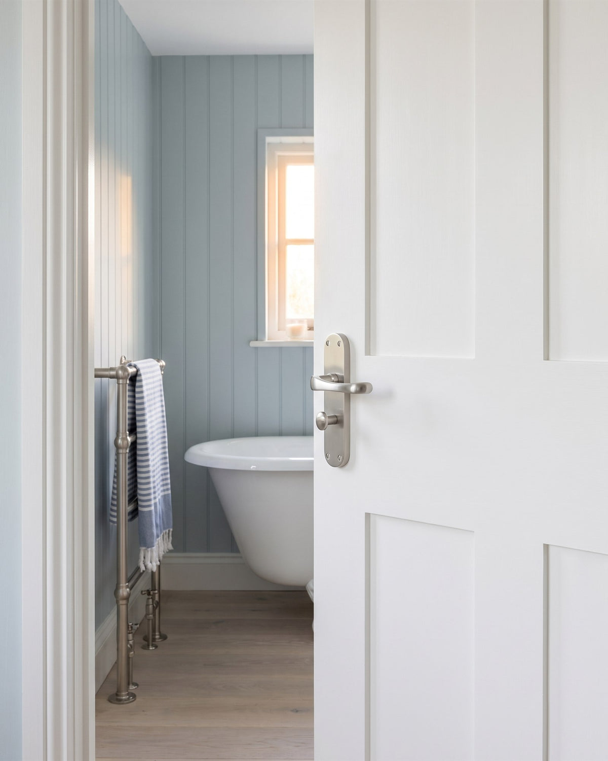 Gloucester satin nickel bathroom handle with turn on a white door, opening onto a blue panelled bathroom with a roll-top bath