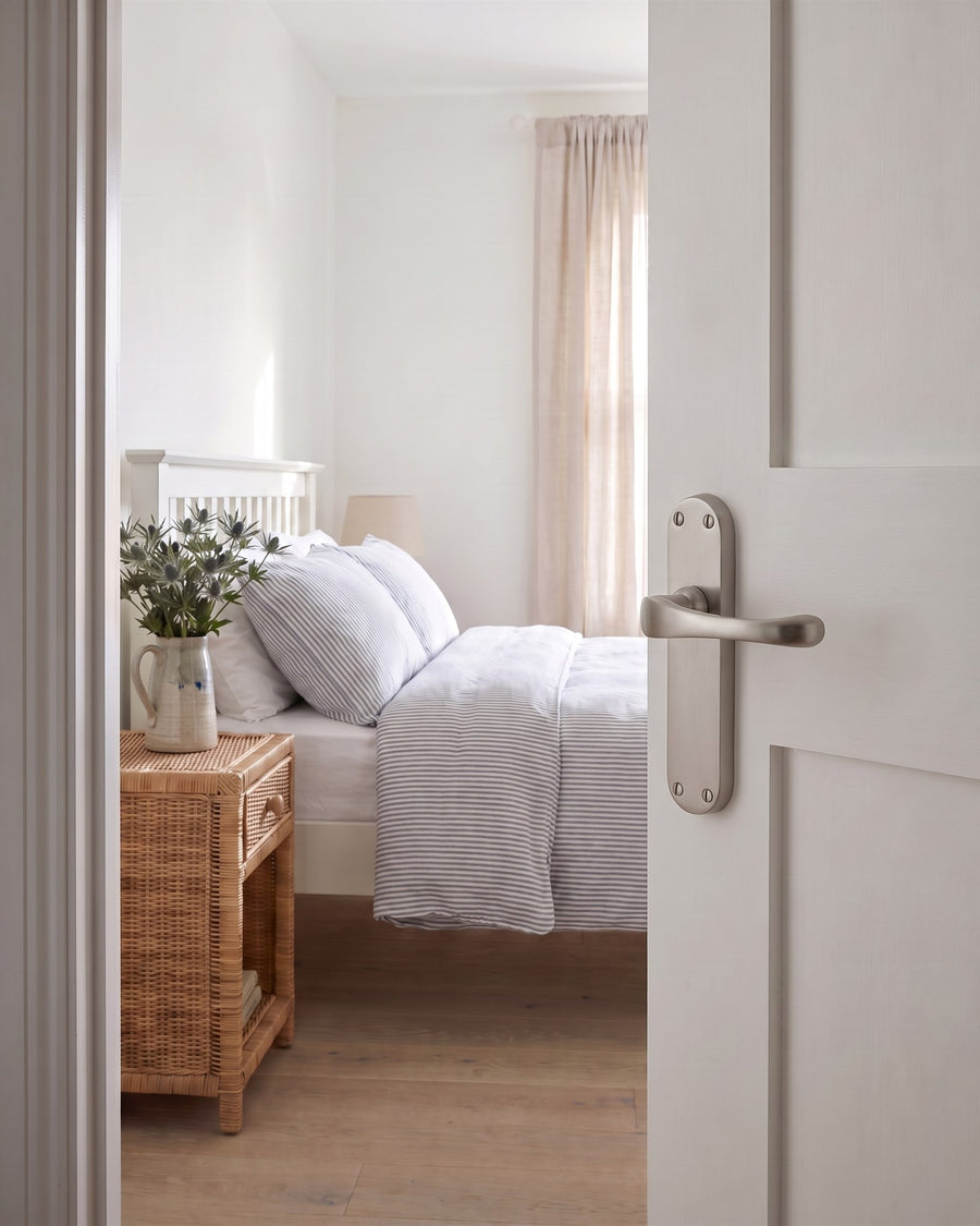 Gloucester satin nickel lever on an open white bedroom door, with ticking-stripe bedding and a rattan bedside table beyond