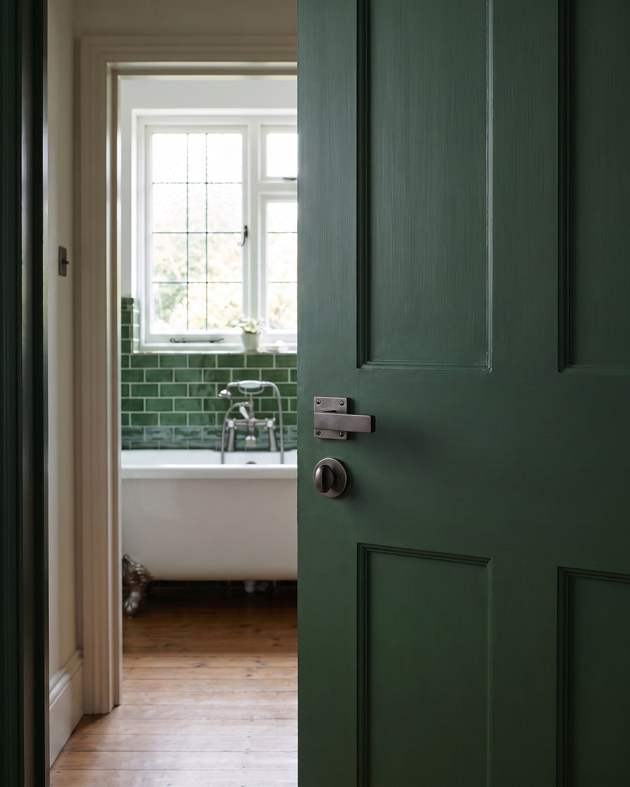 Deep green bathroom door with a rustic pewter lever and round thumb turn, open onto green metro tiles and a roll-top bath