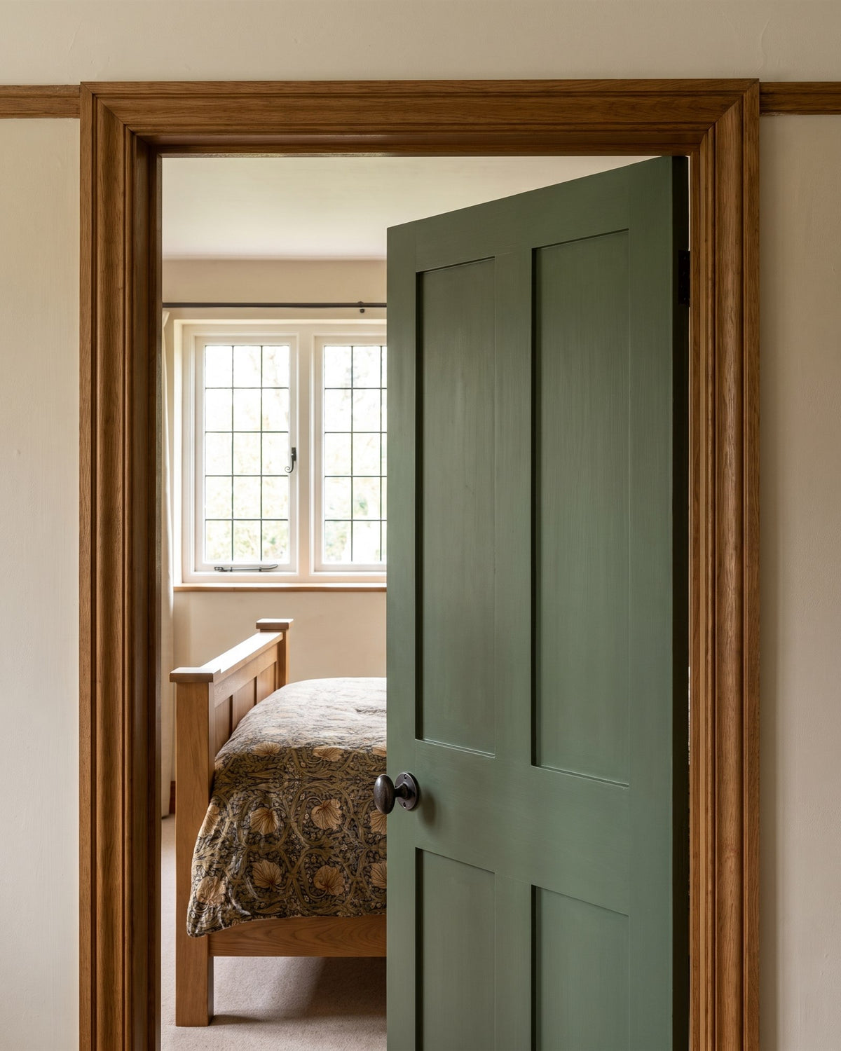 Deep green bedroom door with a rustic pewter oval knob, opening onto an oak bed with a William Morris print bedspread