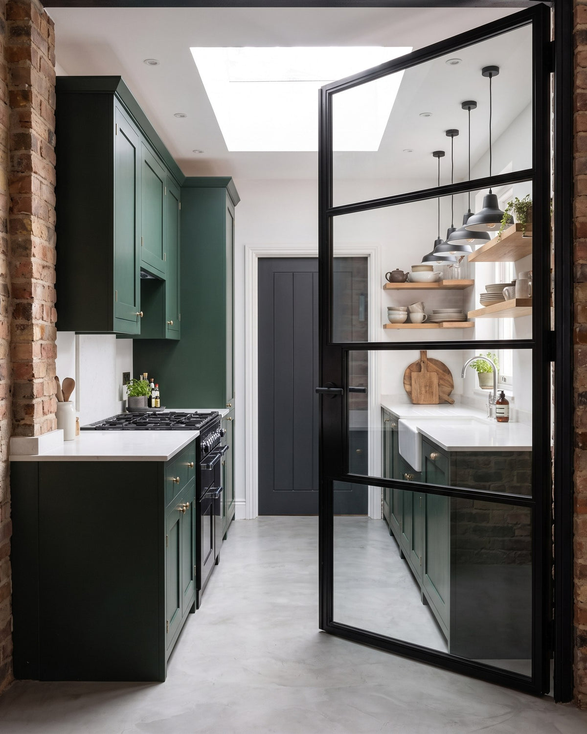 Green shaker kitchen with exposed brick, a range cooker and open oak shelves, seen through an open black-framed glazed door