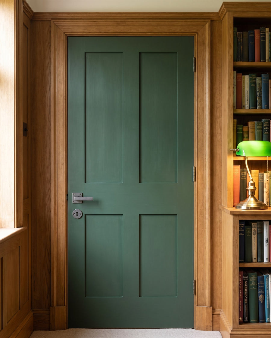 Deep green study door with a rustic pewter lever on square rose and round key escutcheon, beside oak bookshelves and a green banker's lamp