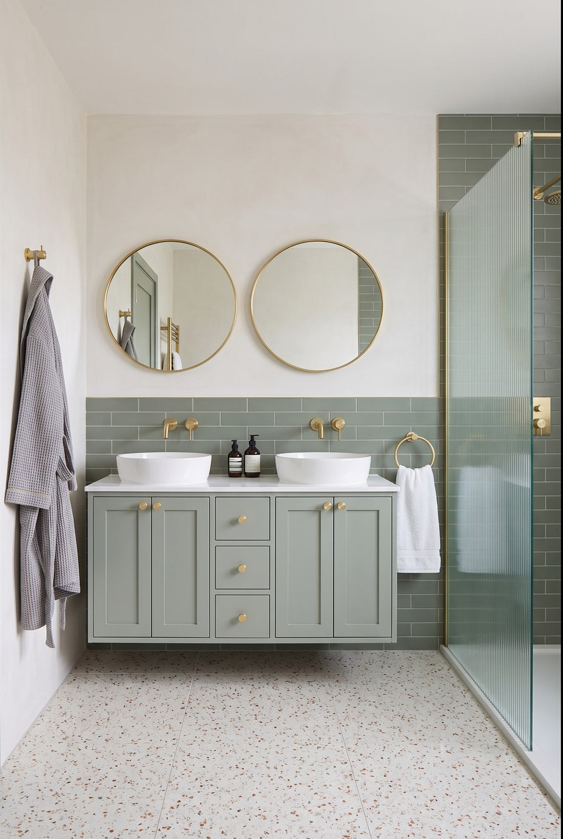 Hotel-style bathroom with a green-grey double vanity fitted with satin brass knurled knobs, two round brass mirrors, a satin brass towel ring, a knurled robe hook and a ribbed glass shower