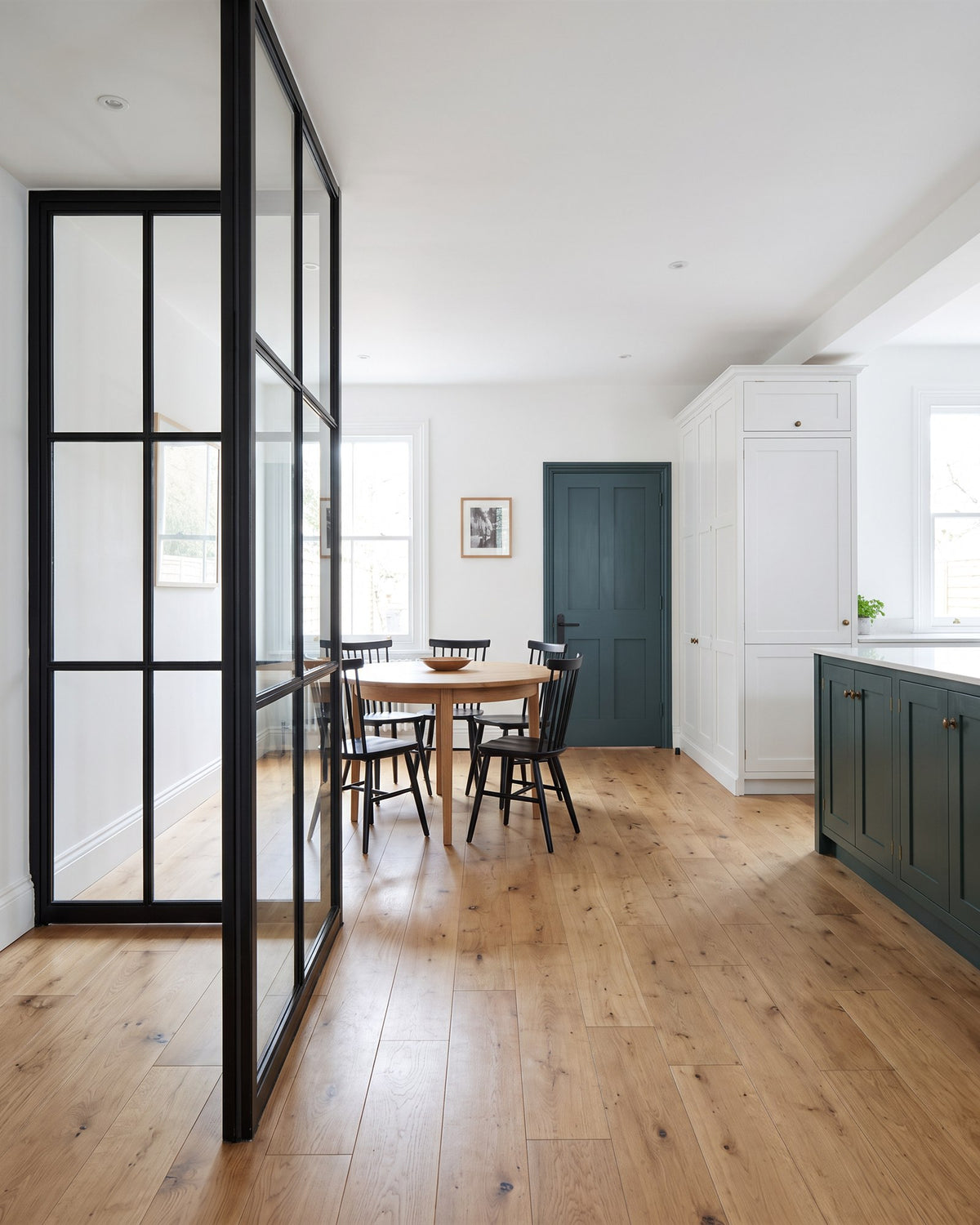 Kitchen extension with an oiled oak floor, black steel glazed screen, round oak table, white kitchen and a deep green island, a green panelled door at the back