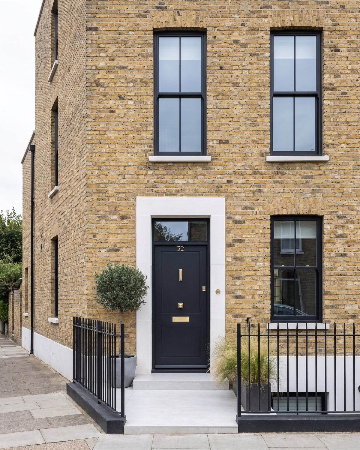 Yellow London stock brick townhouse with black-framed windows, black railings, a clipped olive tree and a near-black front door with satin brass fittings in a white stone surround