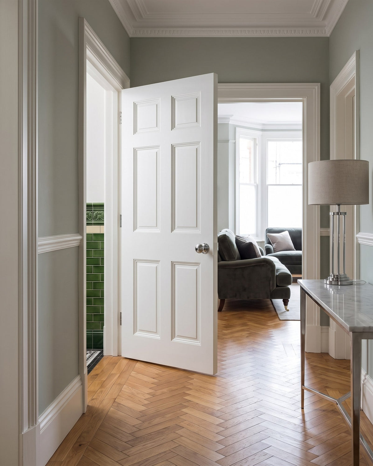 Mansion flat hallway with herringbone oak floor and grey-green walls, a white six-panel door with a polished chrome reeded knob open to a bay-windowed sitting room