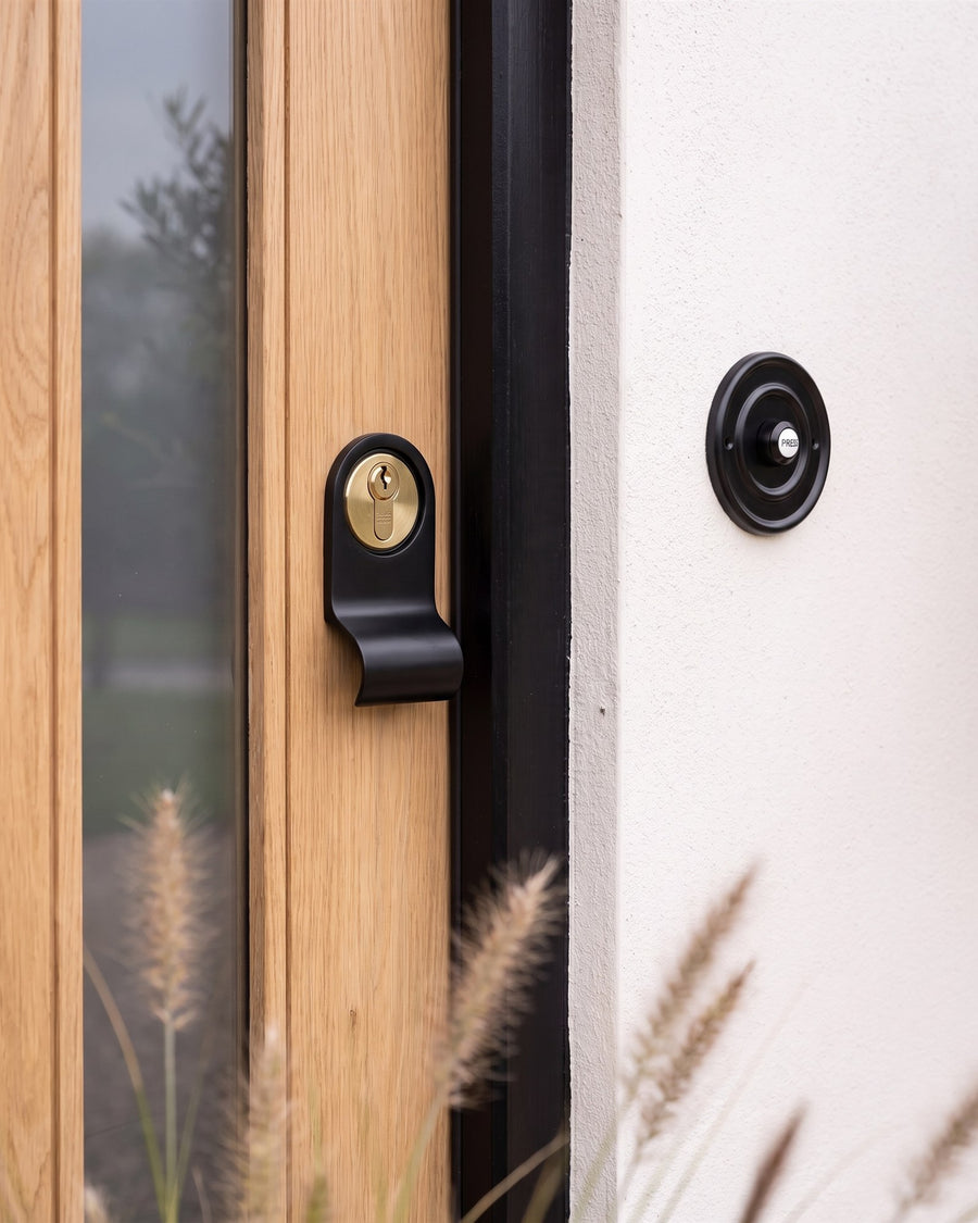 Matt black cylinder pull with a brass euro cylinder on an oak door, beside a round matt black bell push on white render
