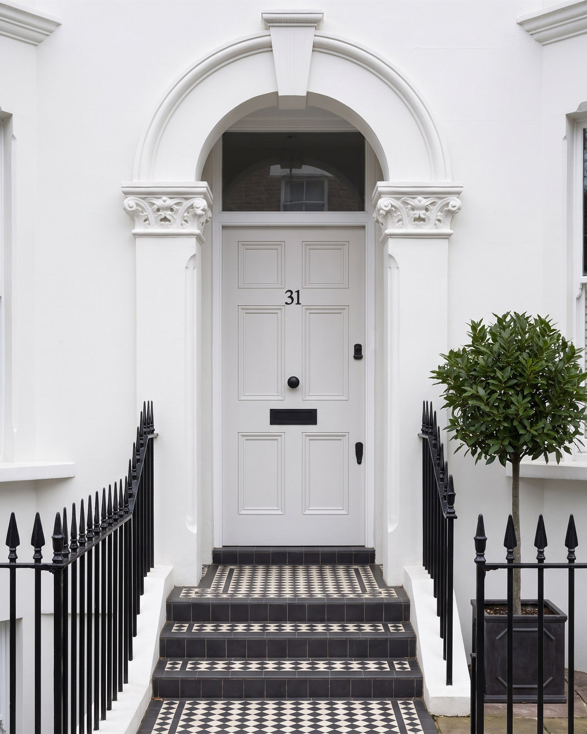 Pale grey-white six-panel front door with matt black number 31, letter plate, knob and cylinder pull under a white stucco arch, tiled steps and black railings