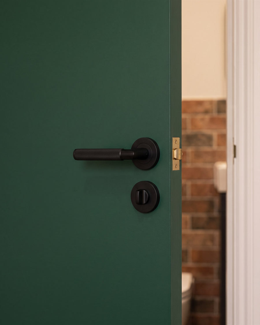 Matt black knurled lever and easy turn on a deep green cloakroom door with brick-effect tiles beyond
