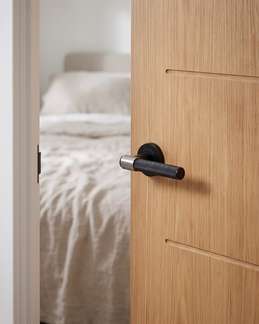 Matt black knurled T-bar lever with a polished collar on an oak veneer bedroom door, linen bedding beyond