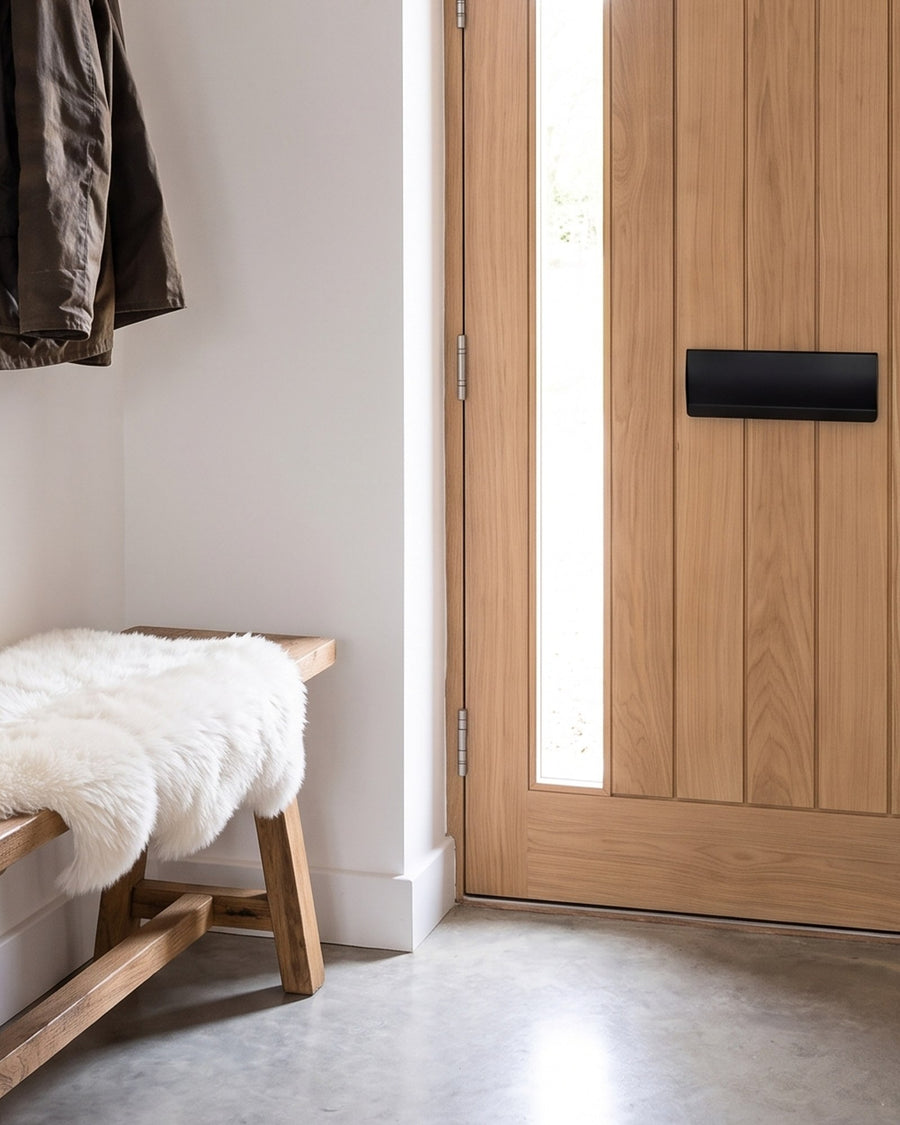 Matt black letter box tidy on the inside of an oak front door beside an oak bench with a sheepskin on a polished concrete floor